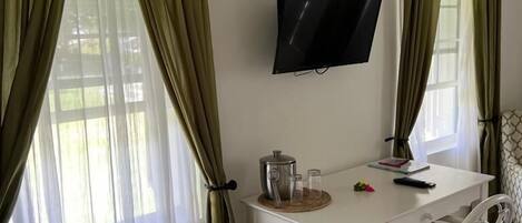 Una habitación con una mesa blanca, un televisor montado en la pared y un ventanal con cortinas verdes.