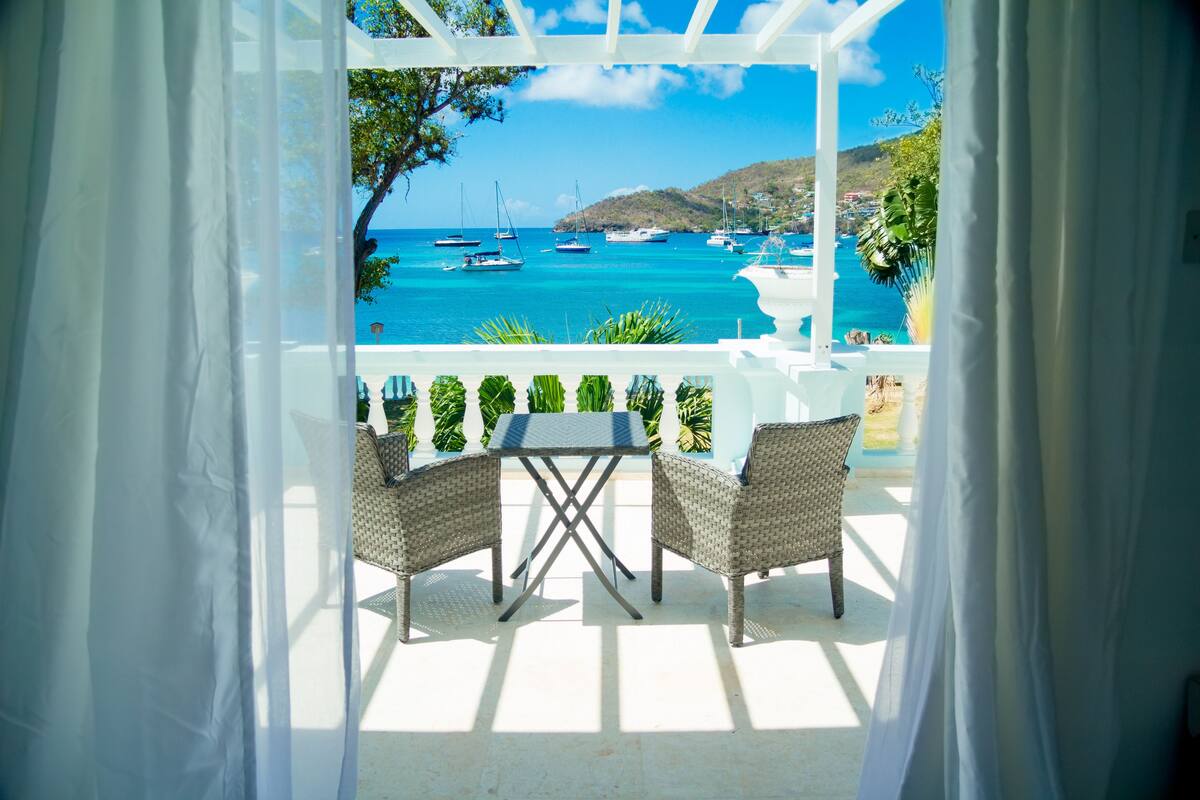 Un espacio de estar al aire libre cubierto con sillas de mimbre y una mesa, con vistas a un mar azul y despejado con barcos y un paisaje montañoso.