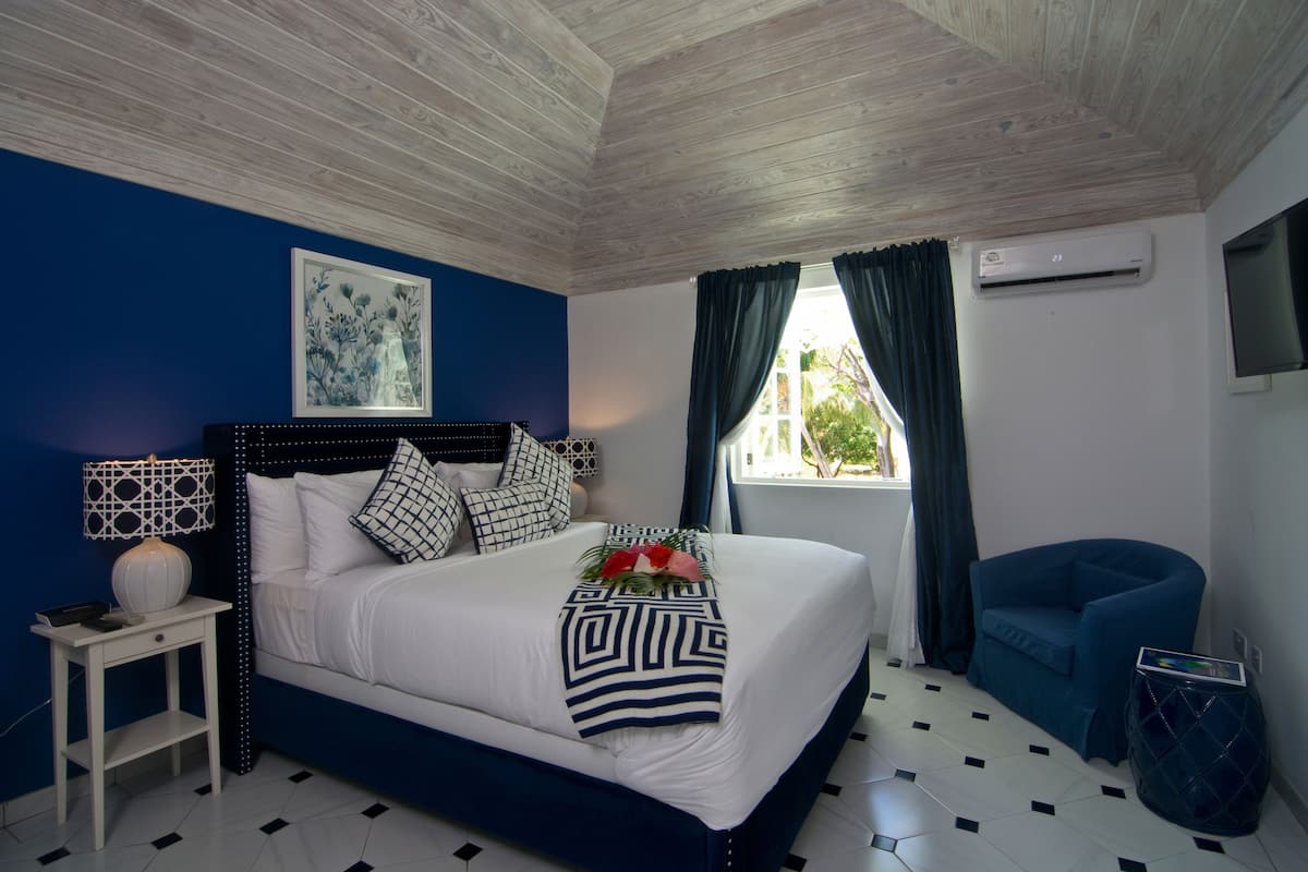 Un dormitorio con cama, mesita de noche, sillón azul, ventana con cortinas y aire acondicionado de pared.