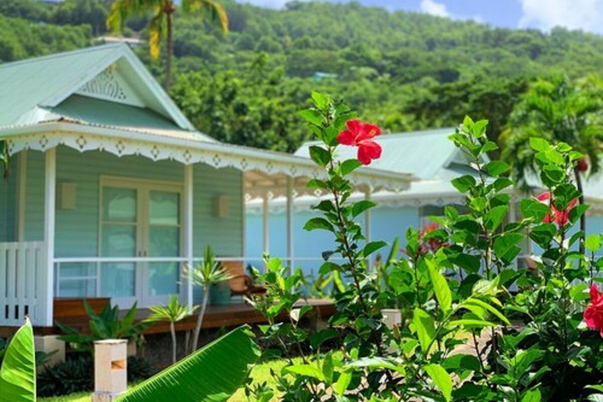 Una casa con techo verde y detalles blancos, rodeada de abundante follaje y un paisaje tropical.