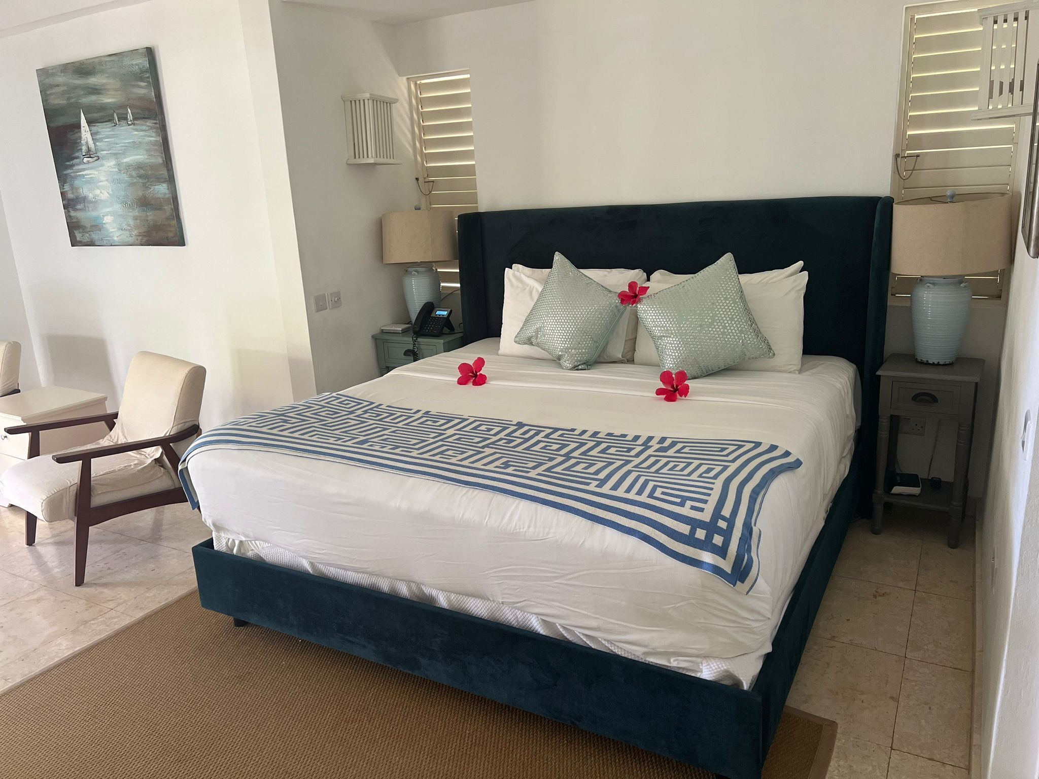 Un cuarto de hotel ordenado con una cama grande, una silla, una mesita de noche y un cuadro colgado en la pared.