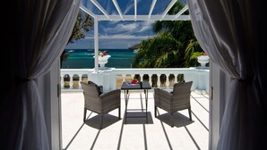 A covered outdoor seating area with two wicker chairs and a table, overlooking a turquoise sea with boats and palm trees.