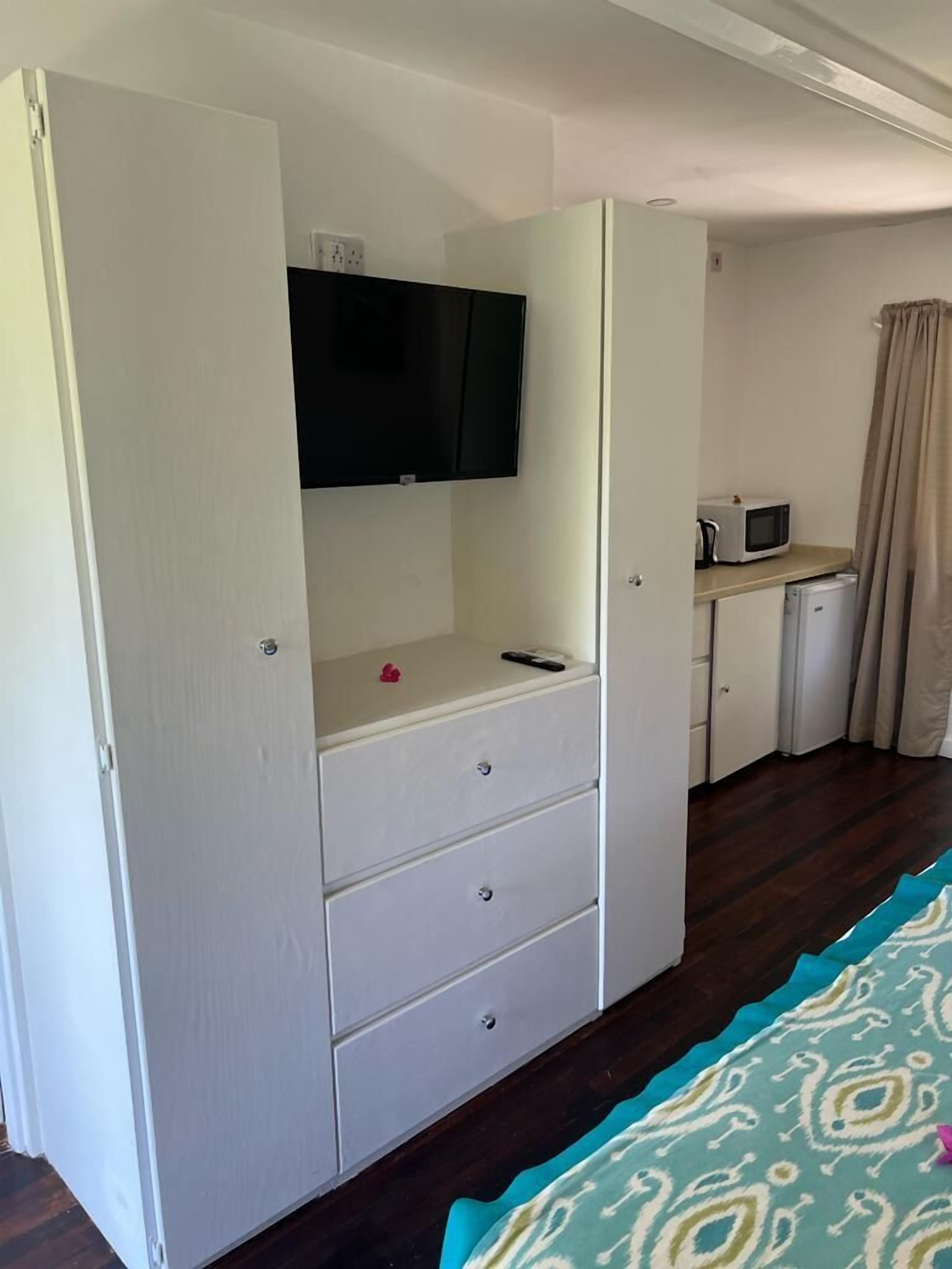 Ein kompaktes Zimmer mit einem weißen Kleiderschrank, einem Flachbildfernseher über einer Kommode, einer kleinen Küchenzeile inklusive Mikrowelle und einem gemusterten Teppichboden.