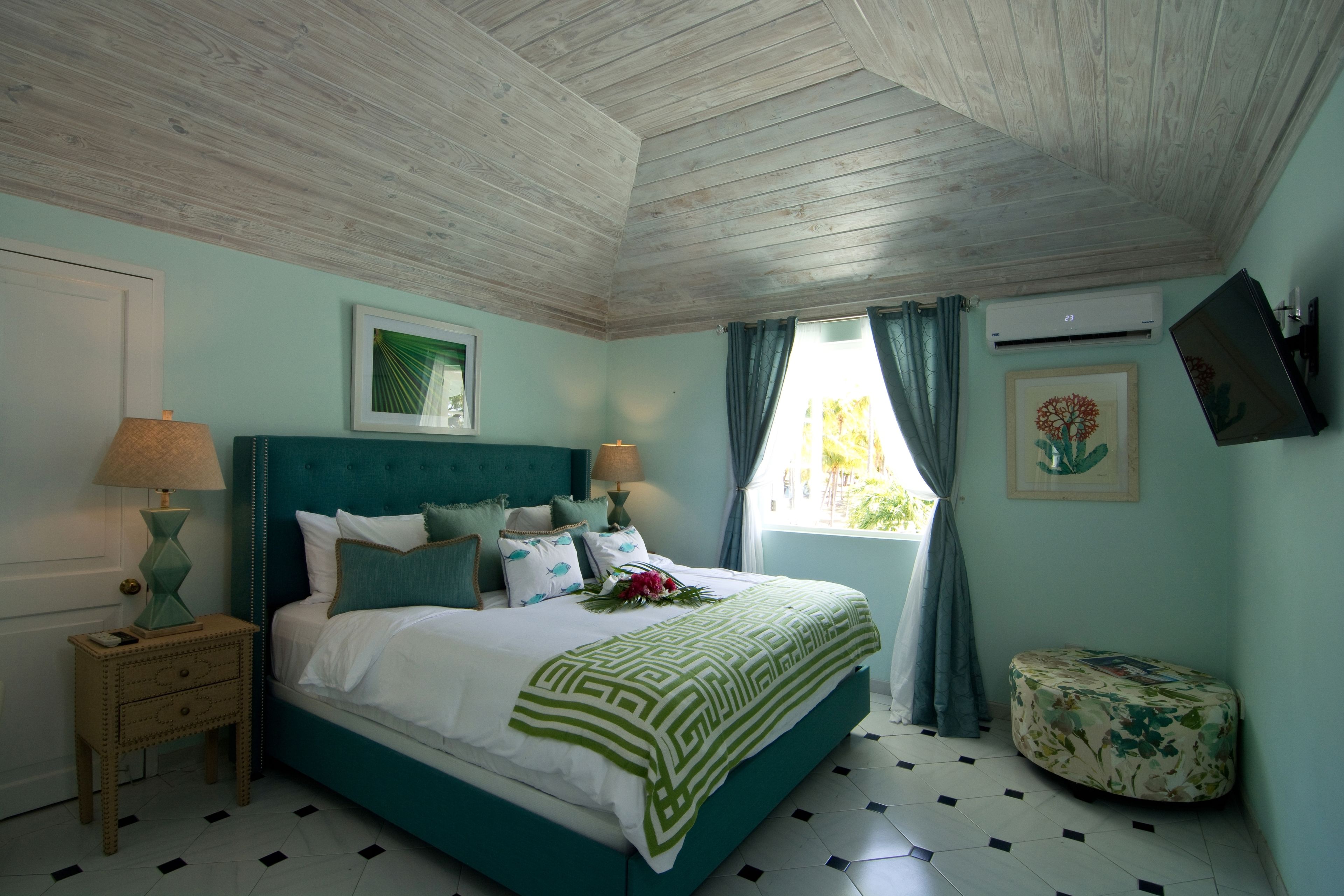 Una camera da letto con un letto color petrolio, soffitto in legno, una finestra con tende e un piccolo comodino con una lampada.
