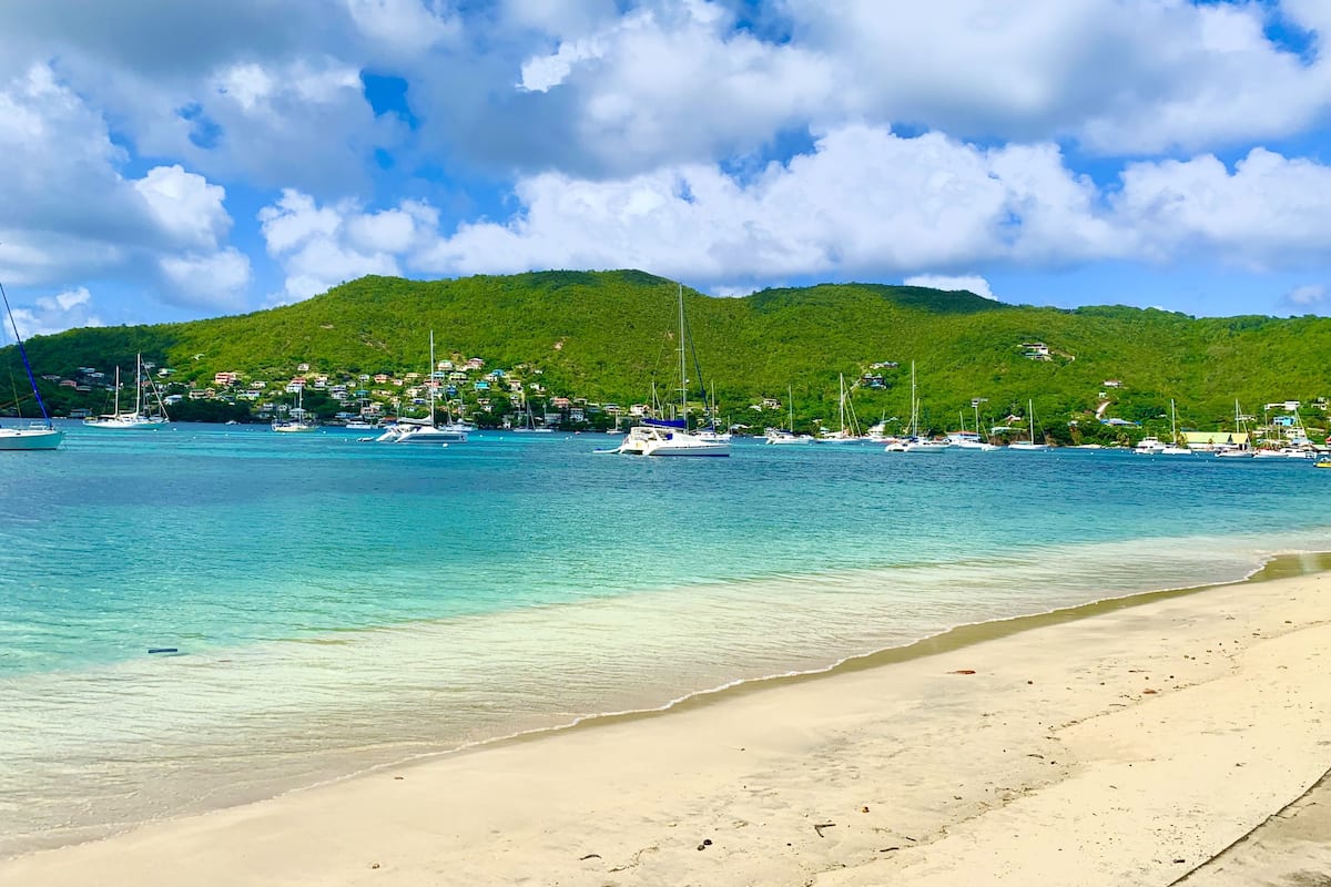Una playa con agua turquesa cristalina, una orilla arenosa y una colina verde al fondo con barcos anclados cerca.