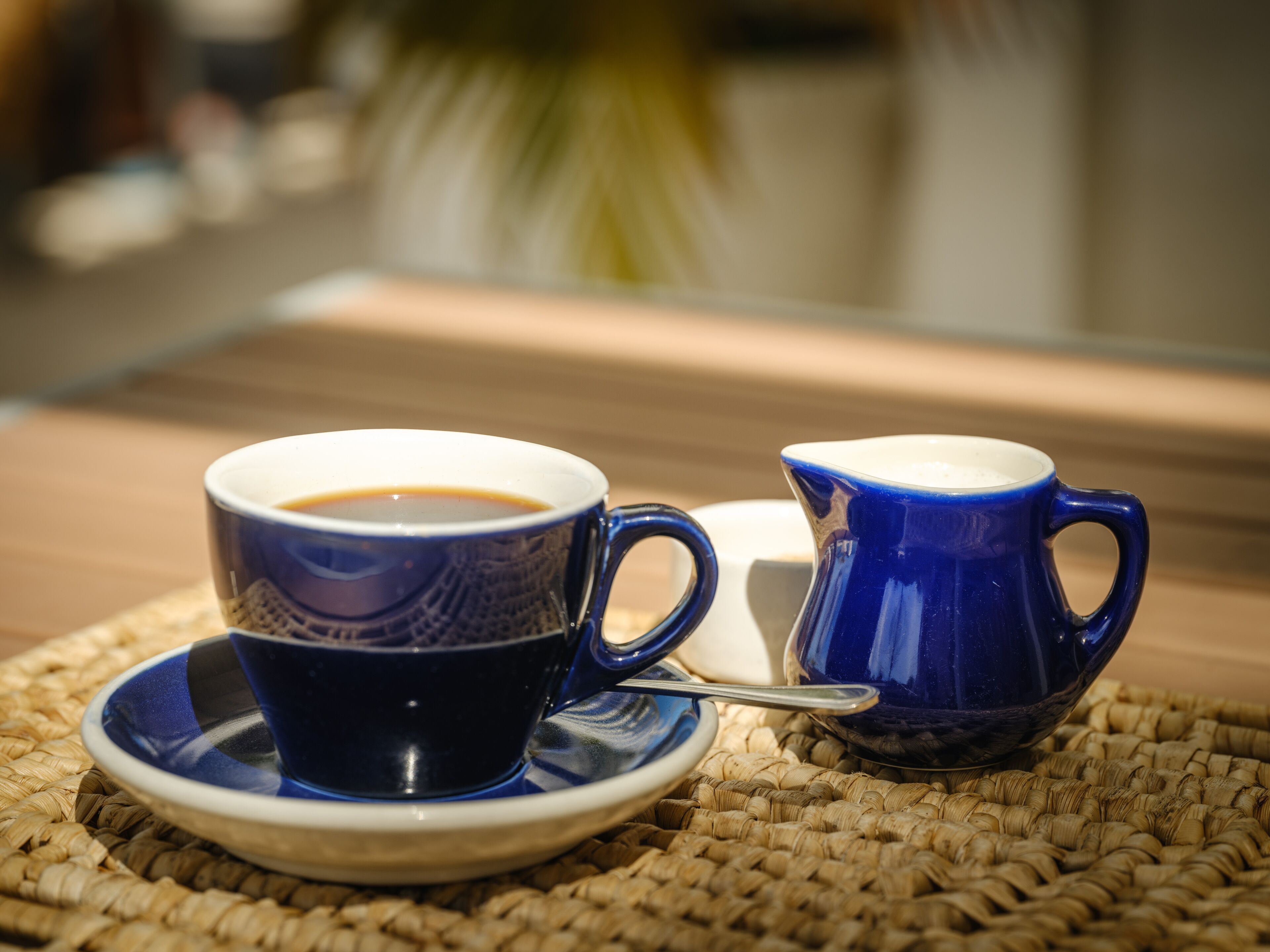 A blue cup of coffee on a woven mat with a saucer and a matching blue cup beside it.