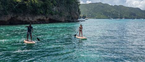 Zwei Personen stehen auf Paddleboards auf ruhiger See, mit einer felsigen Küste und Segelbooten im Hintergrund.