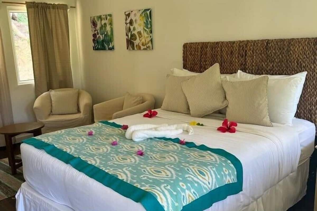 Una cama bien hecha con colcha blanca y azul verdosa, un cabecero marrón y un par de sillones color beige.