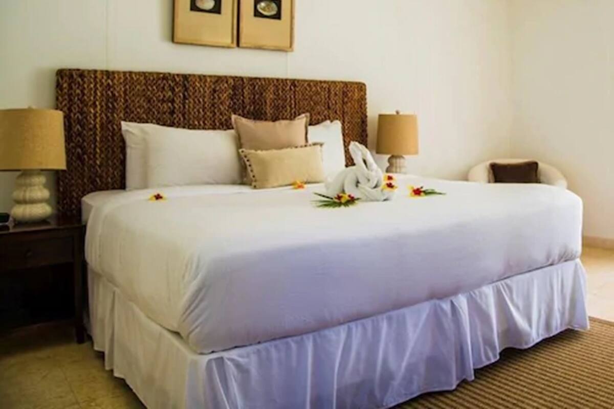 Una cama bien hecha con ropa blanca y un cabecero marrón, dos lámparas de noche y un cuadro enmarcado sobre la cama.