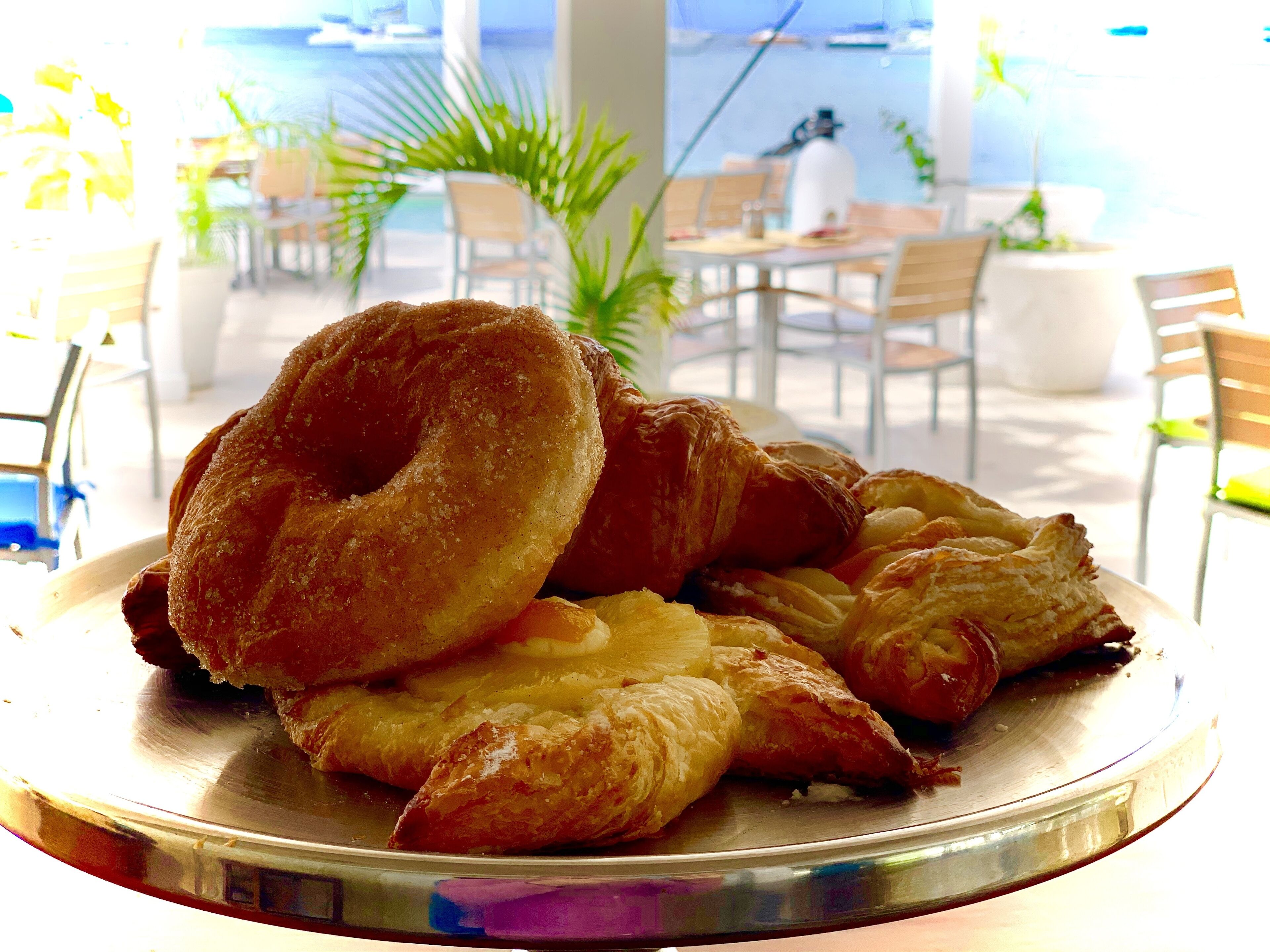 A tray of pastries on a table in a cafe with chairs and a view of the sea.