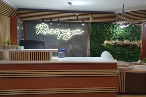 Un área de recepción con diseño moderno, que incluye un mostrador de recepción, un letrero de neón y una pared verde con plantas.