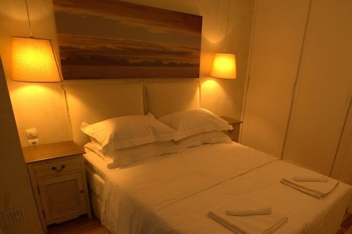 Ein ordentlich bezogenes Bett mit weißen Leinentüchern und Kissen, ein Nachttisch mit Lampe und ein Gemälde über dem Bett.