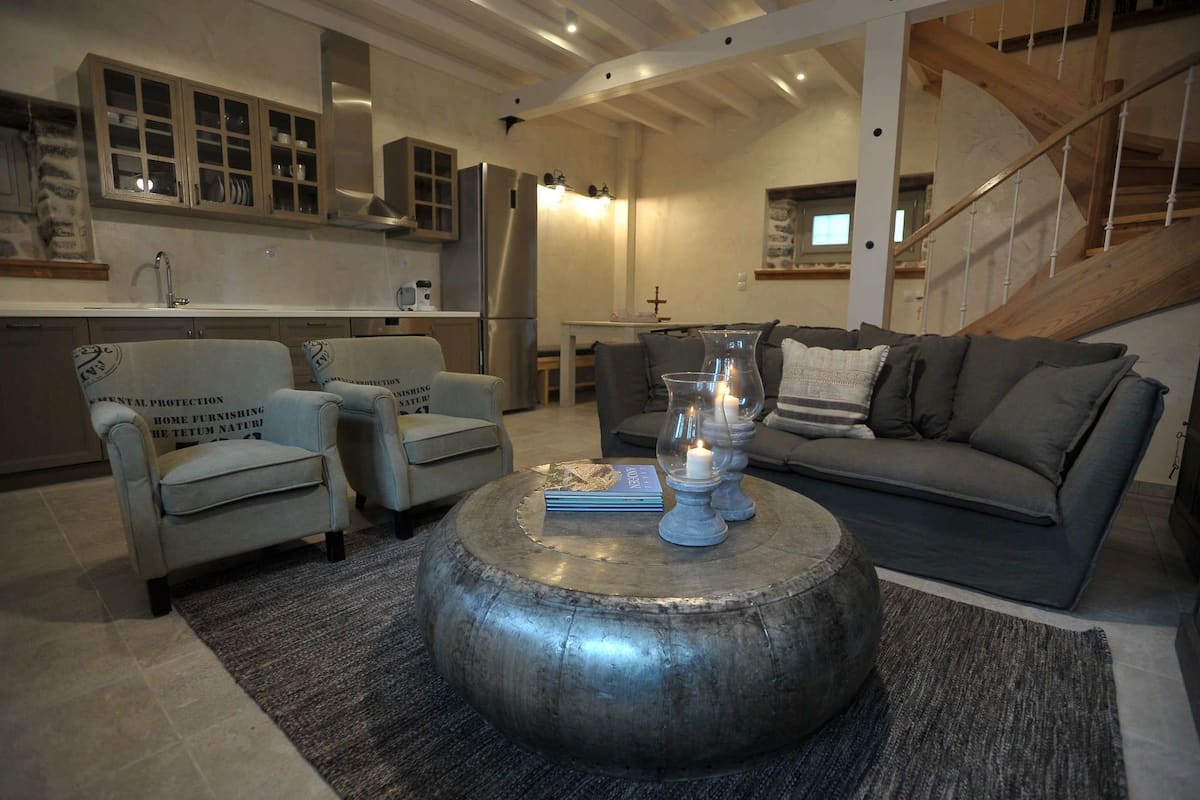 Ein modernes Wohnzimmer mit einem grauen Sofa, zwei Sesseln, einem Couchtisch mit Büchern und Kerzen, und einer Küche im Hintergrund.