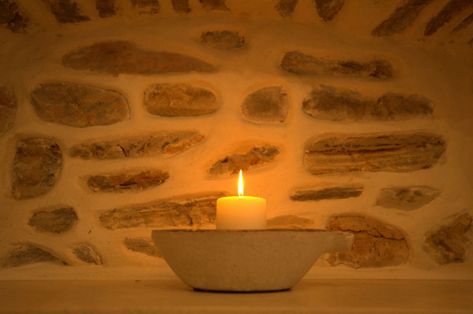 A candle in a stone bowl on a table against a stone wall.