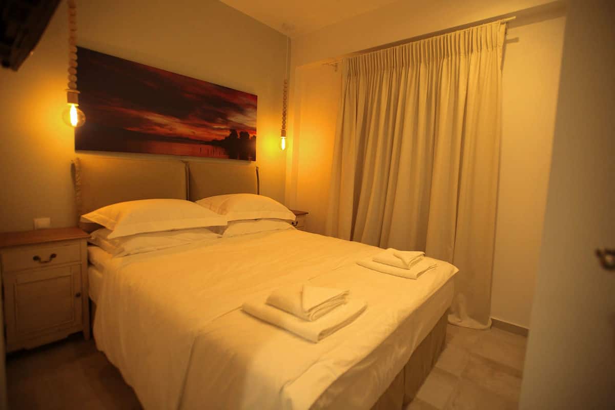 Ein ordentlich gemachtes Bett mit weißen Leinen und Handtüchern, ein Nachttisch mit Lampe und ein Sonnenuntergangsbild an der Wand.