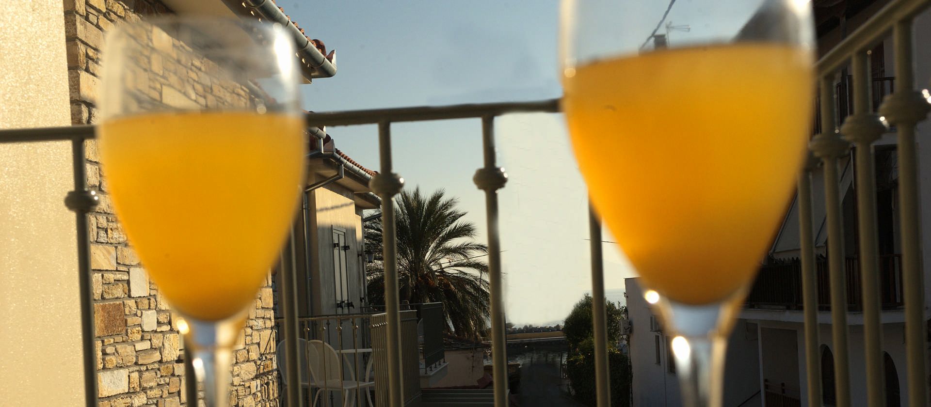 Two glasses of orange beverage on a balcony railing with a view of a stone building and palm trees.
