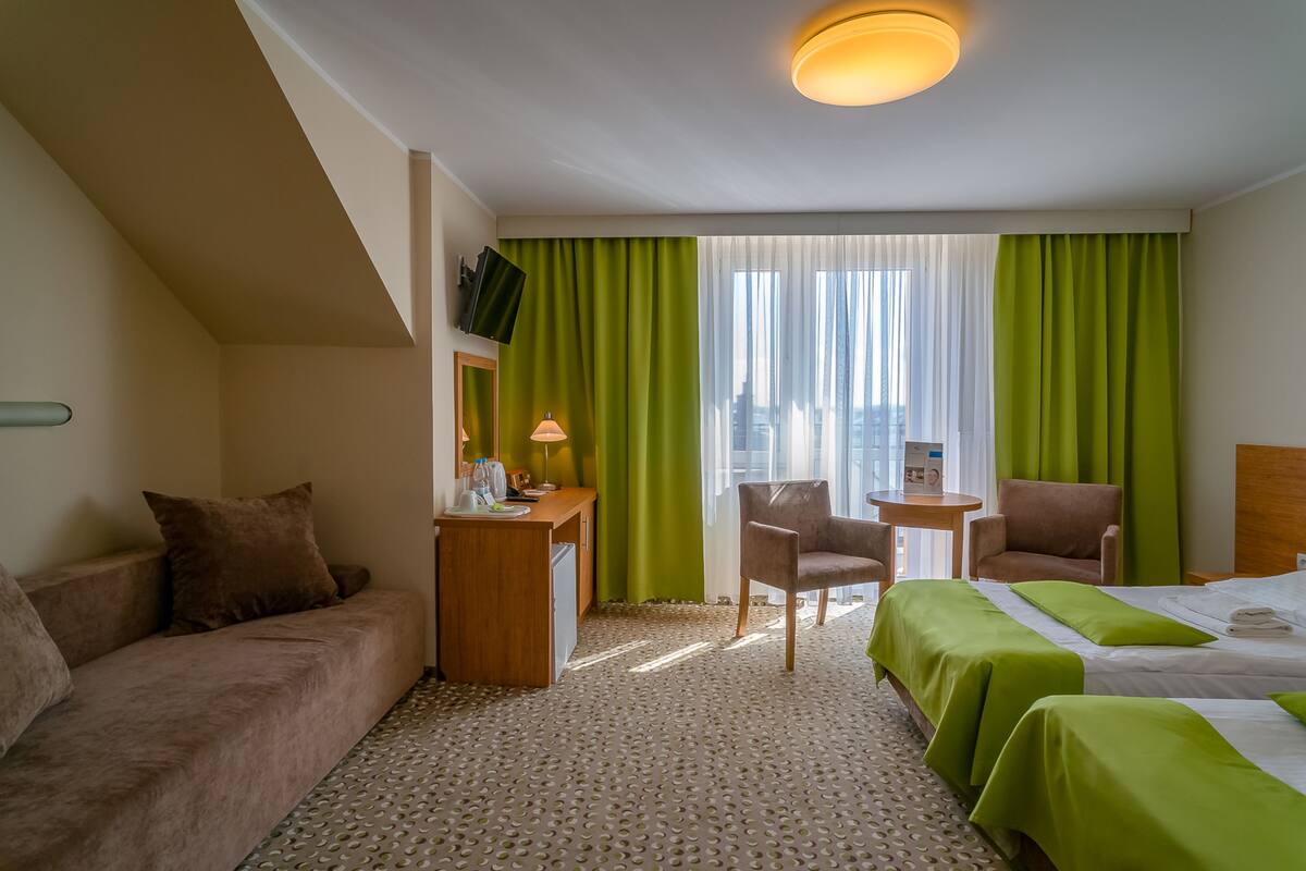 A hotel room with a sofa, two single beds, a desk with a lamp, a chair, and a TV on the wall.