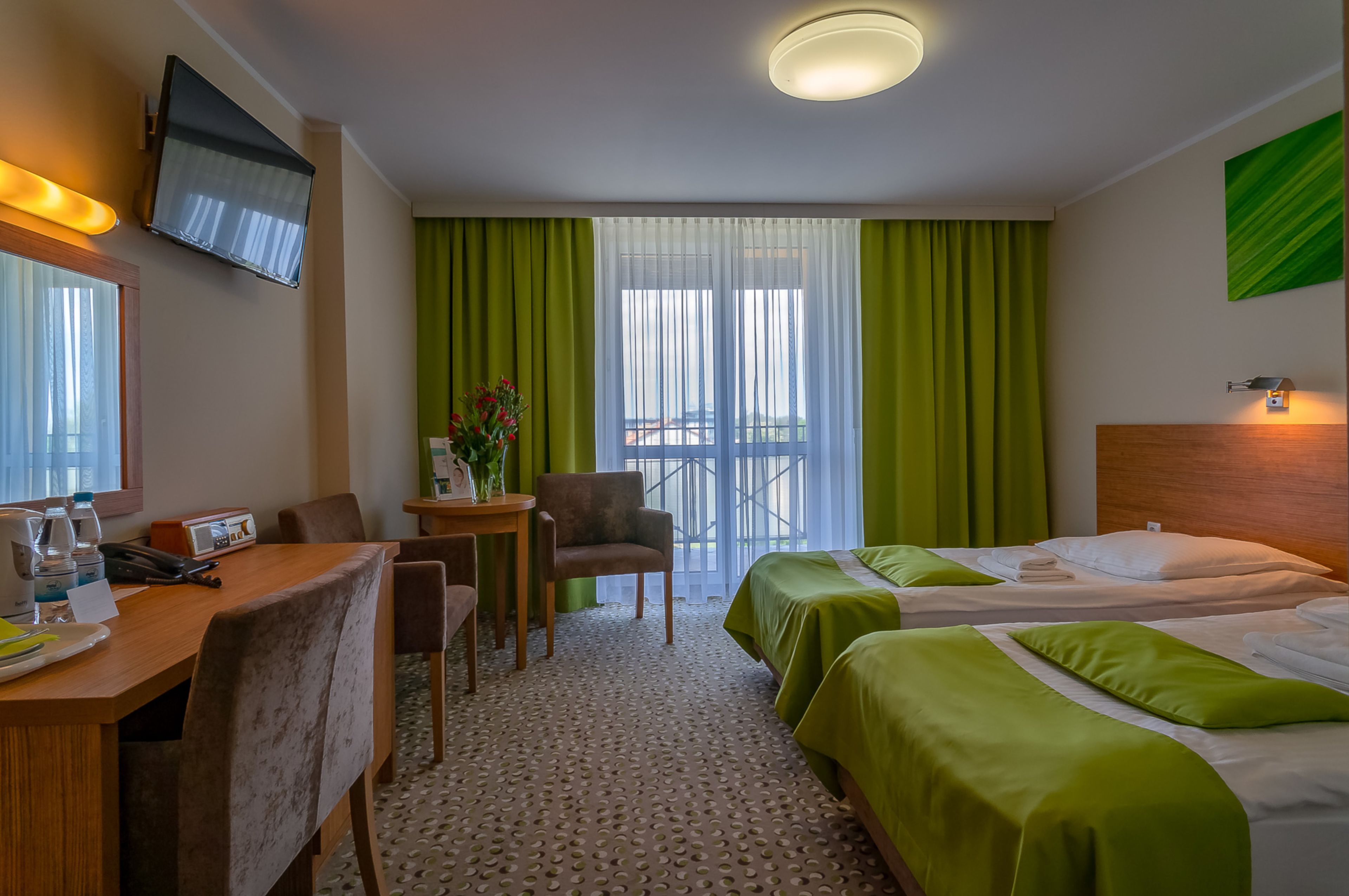 A hotel room with two beds, a desk with a chair, a small table with a plant, and a TV mounted on the wall.