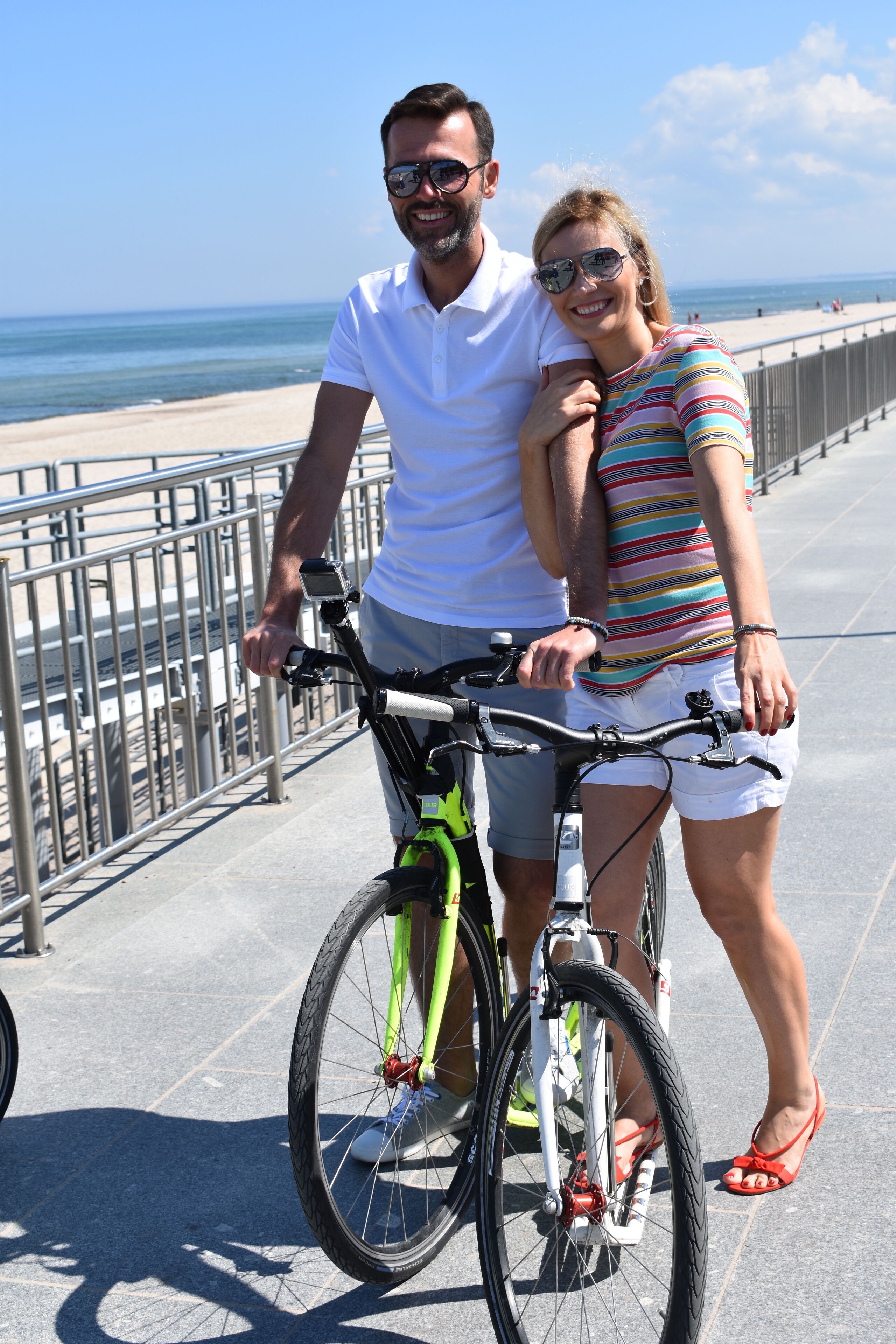 A couple standing next to a bicycle on a boardwalk with a beach in the background.
