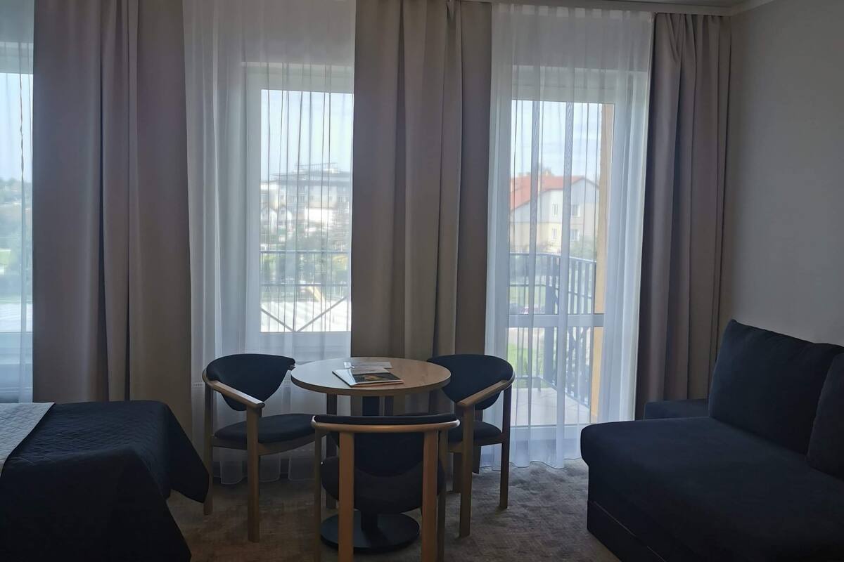 A hotel room with a bed, a round table with chairs, a sofa, and a large window with curtains.