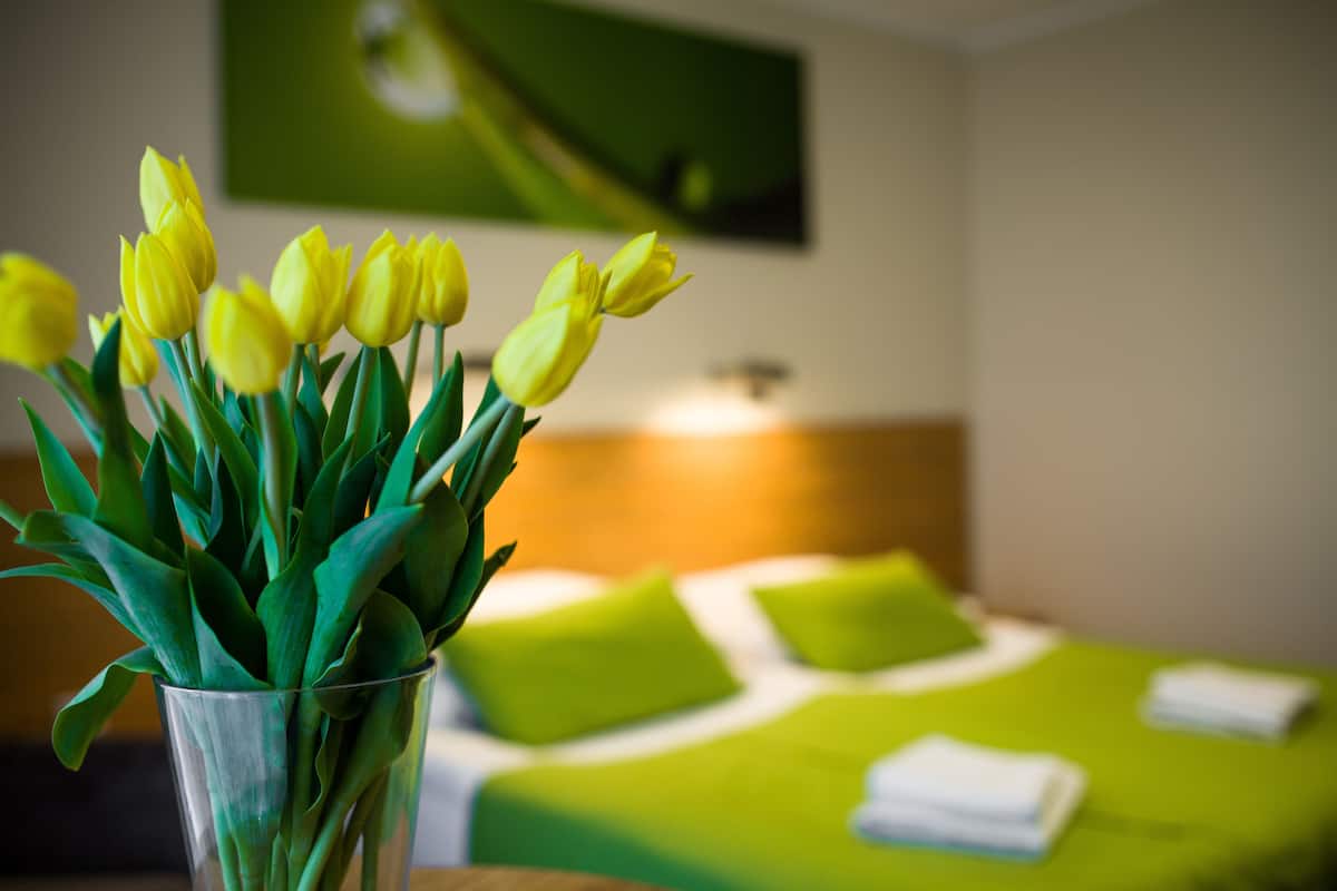 A hotel room with a bed, green bedding, a vase with yellow tulips, and a painting on the wall.