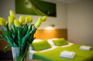 Una habitación de hotel con una cama, ropa de cama verde, un florero con tulipanes amarillos y un cuadro en la pared.