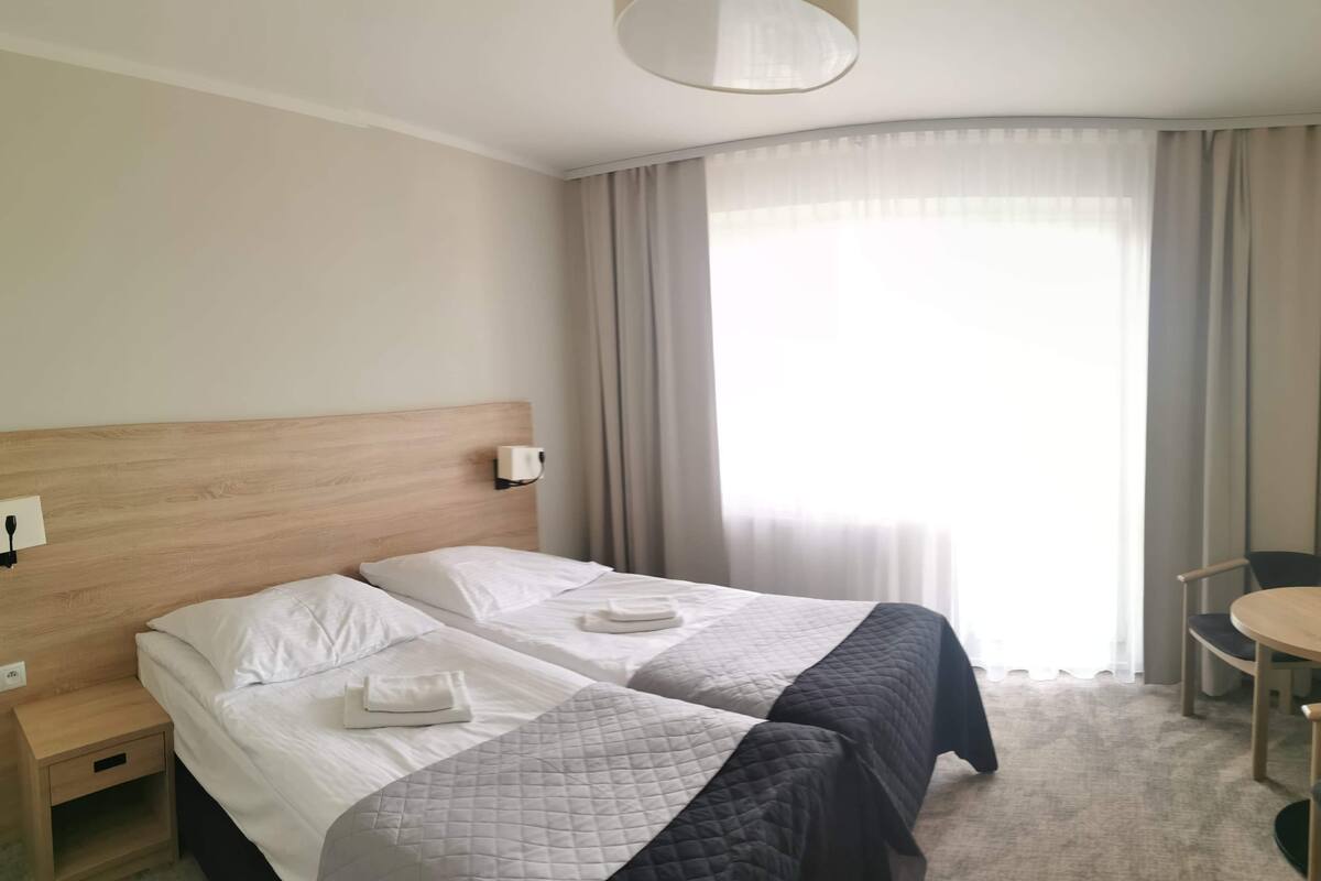 A hotel room with two beds, a wooden headboard, a bedside table, a chair, a small table, and a large window with curtains.
