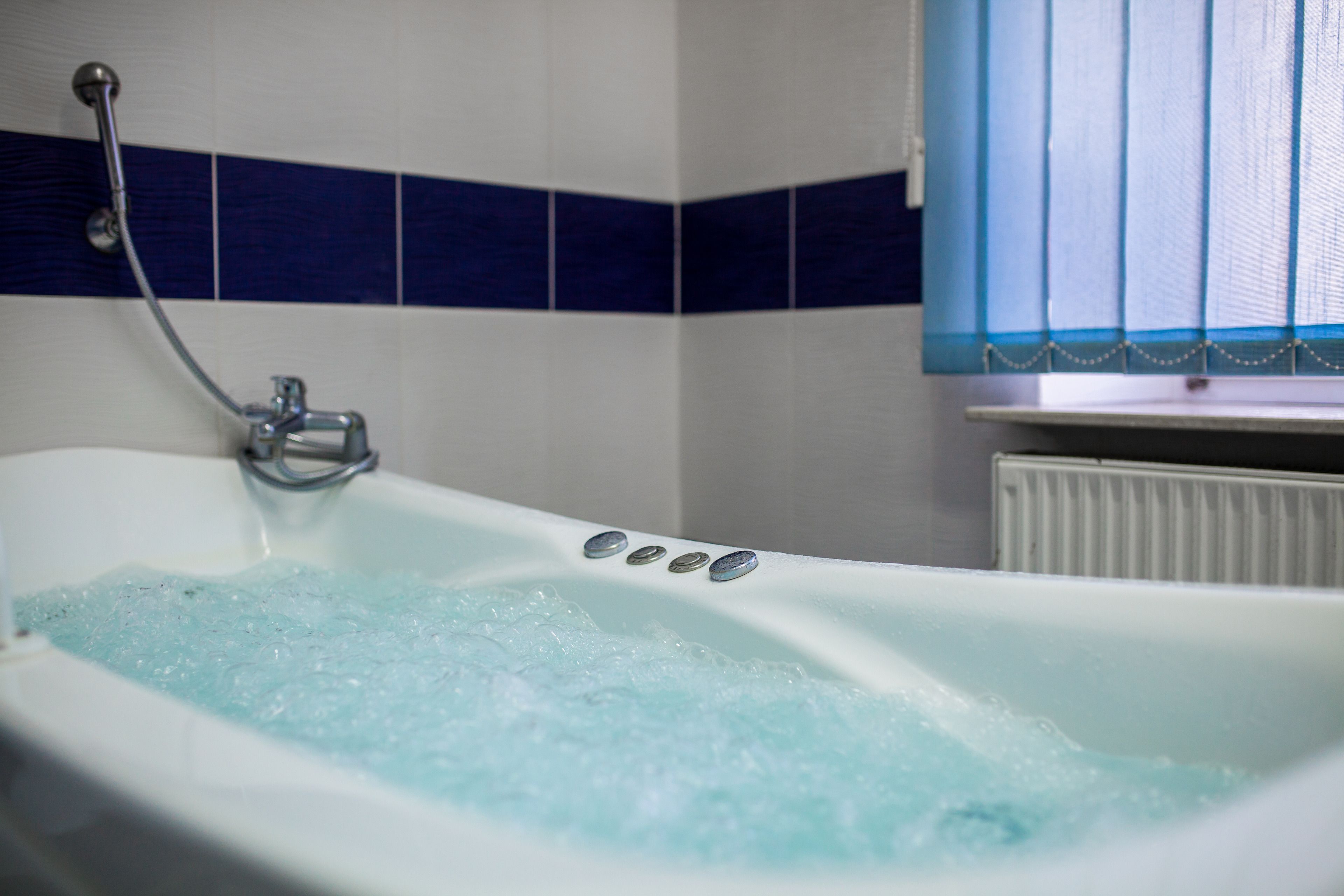 A bathtub with bubbles, a showerhead, and a window with blinds.