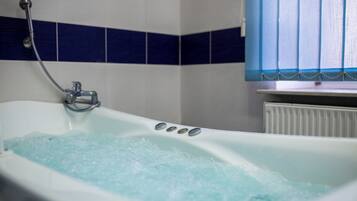 A bathtub with bubbles, a showerhead, and a window with blinds.
