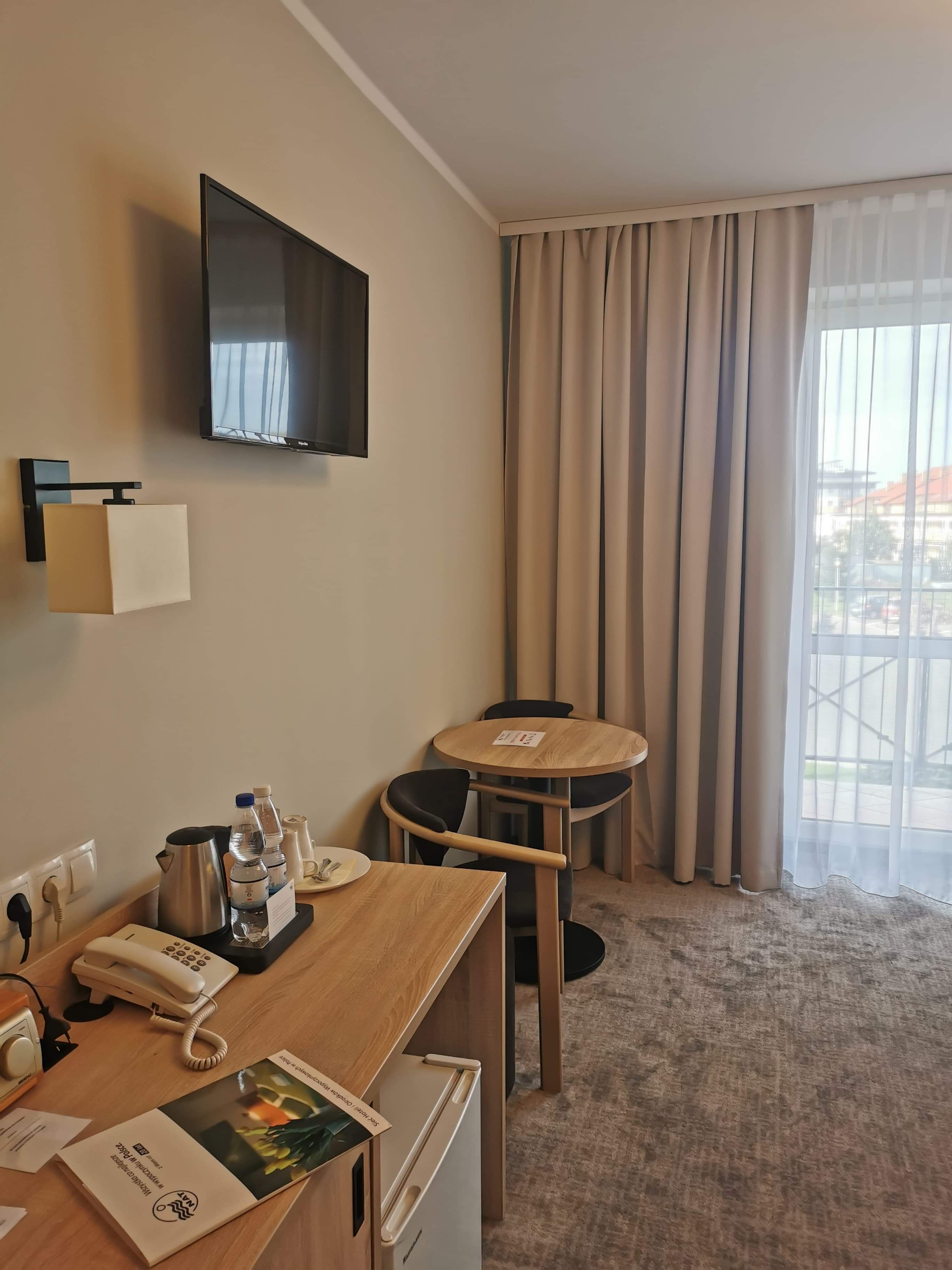 Une chambre d’hôtel avec un bureau en bois, un téléviseur à écran plat fixé au mur, une petite table ronde avec des chaises, une grande fenêtre avec des rideaux et un sol recouvert de moquette.