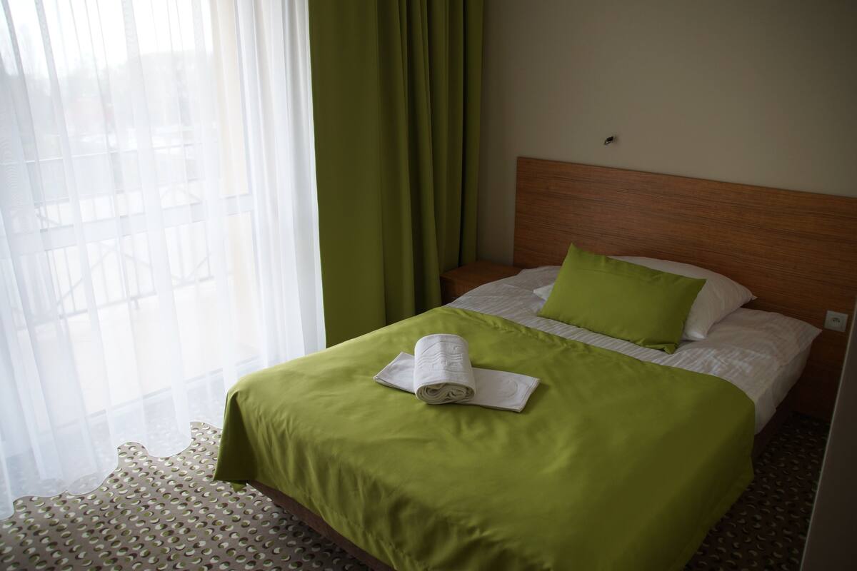 A hotel room with a neatly made bed, green curtains, and a wooden headboard.