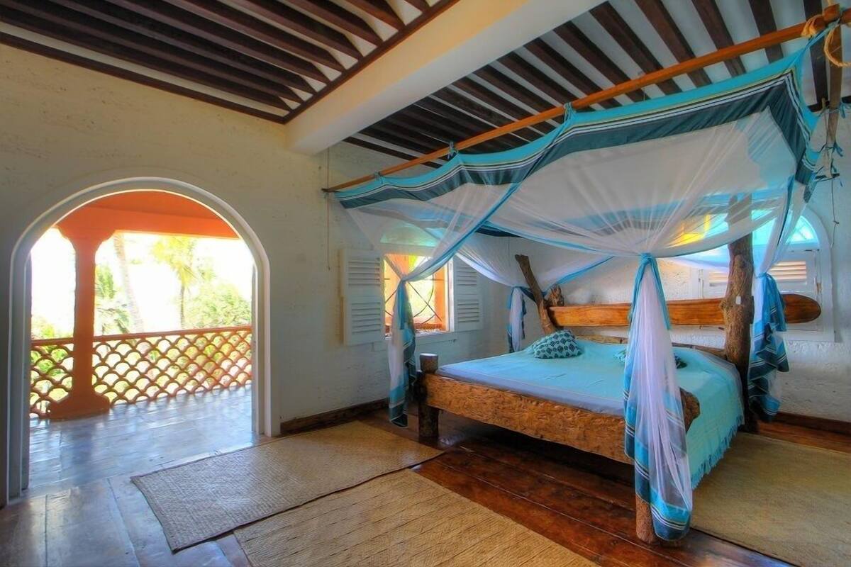 A room with a bed under a canopy, wooden flooring, a view through an archway, and a wooden ceiling.