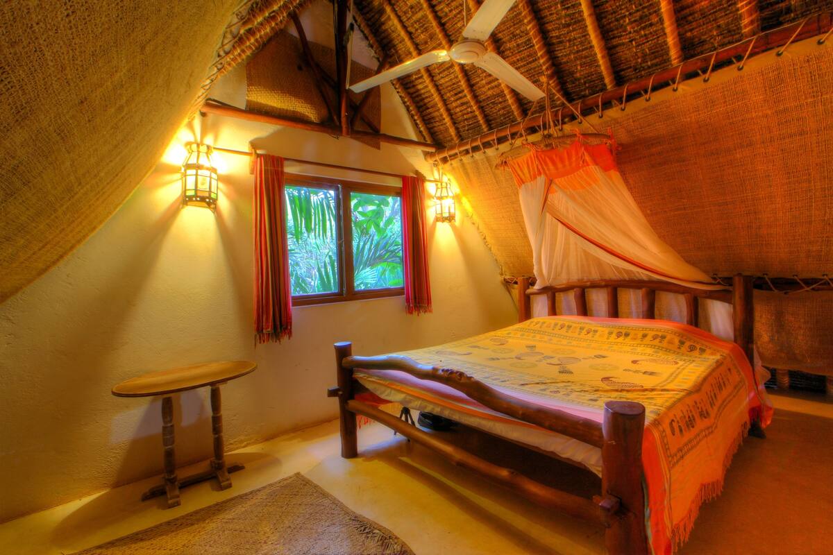 A room with a bed, a small table, a window with curtains, and a ceiling fan.