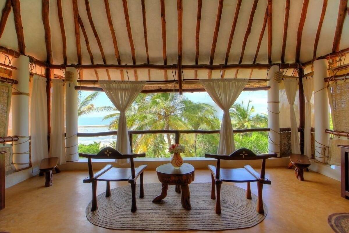 A room with a wooden ceiling, large windows with white curtains, a round table with chairs, and a view of palm trees and the sea.