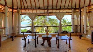 Una habitación con techo de madera, ventanas amplias con cortinas blancas, una mesa redonda con sillas y vistas a palmeras y al mar.