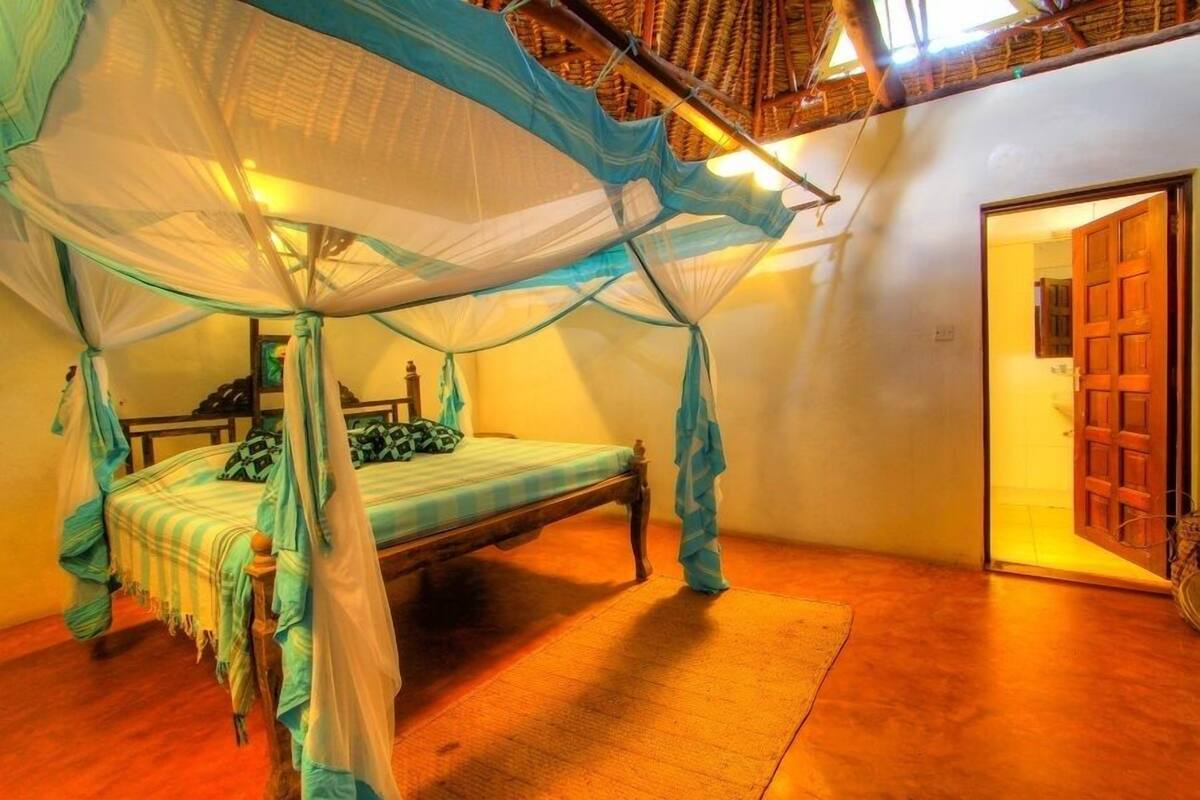 A room with a bed under a mosquito net, wooden flooring, and a door leading to another room.
