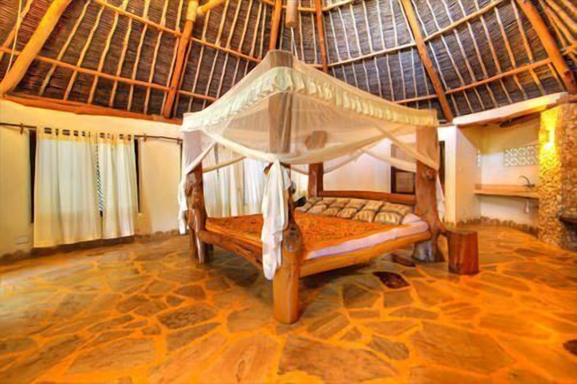 A room with a bed under a thatched roof, stone flooring, and a window with curtains.