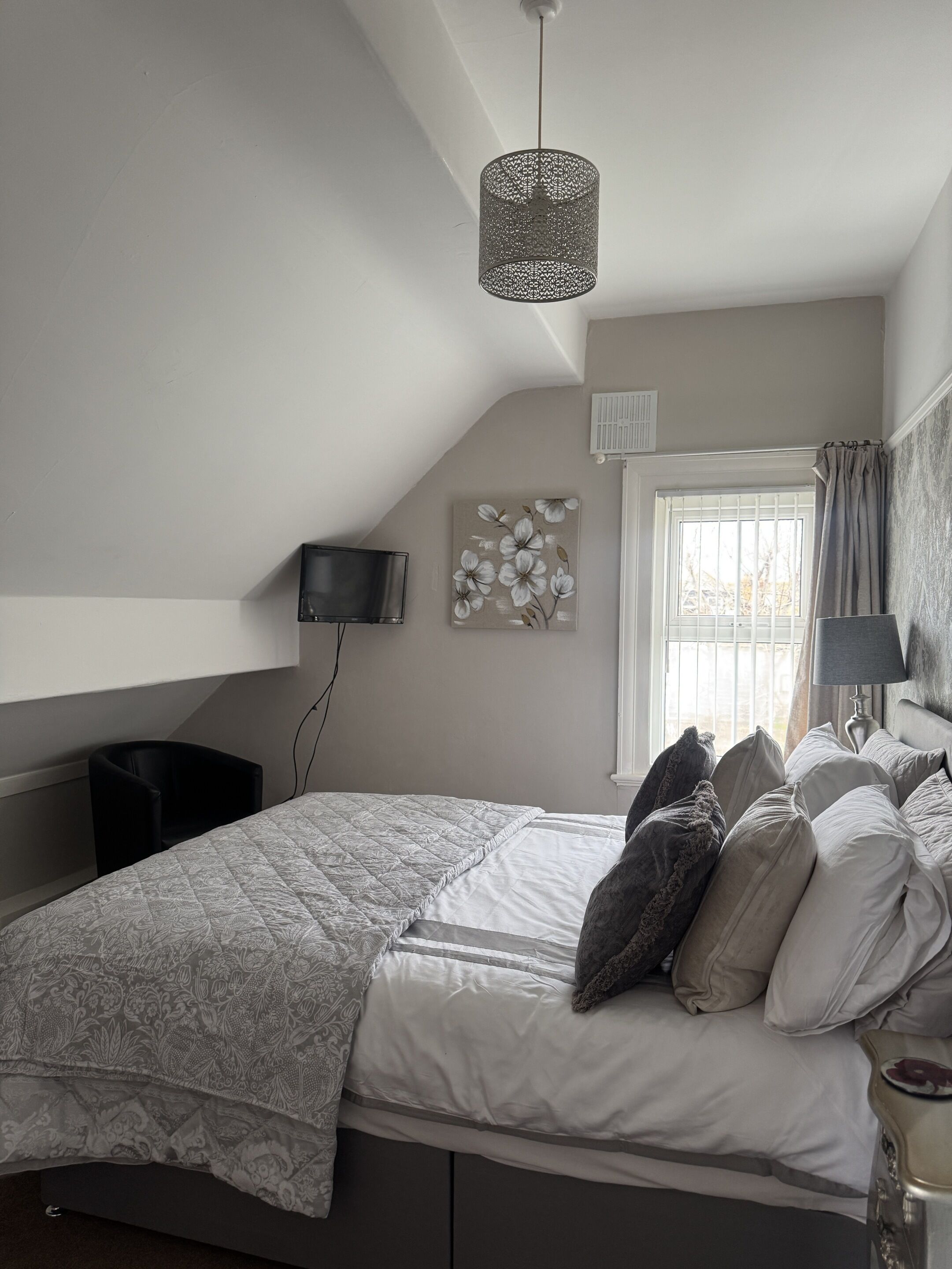 Un dormitorio con una cama grande, un televisor montado en la pared, una lámpara de techo y una ventana con cortinas.