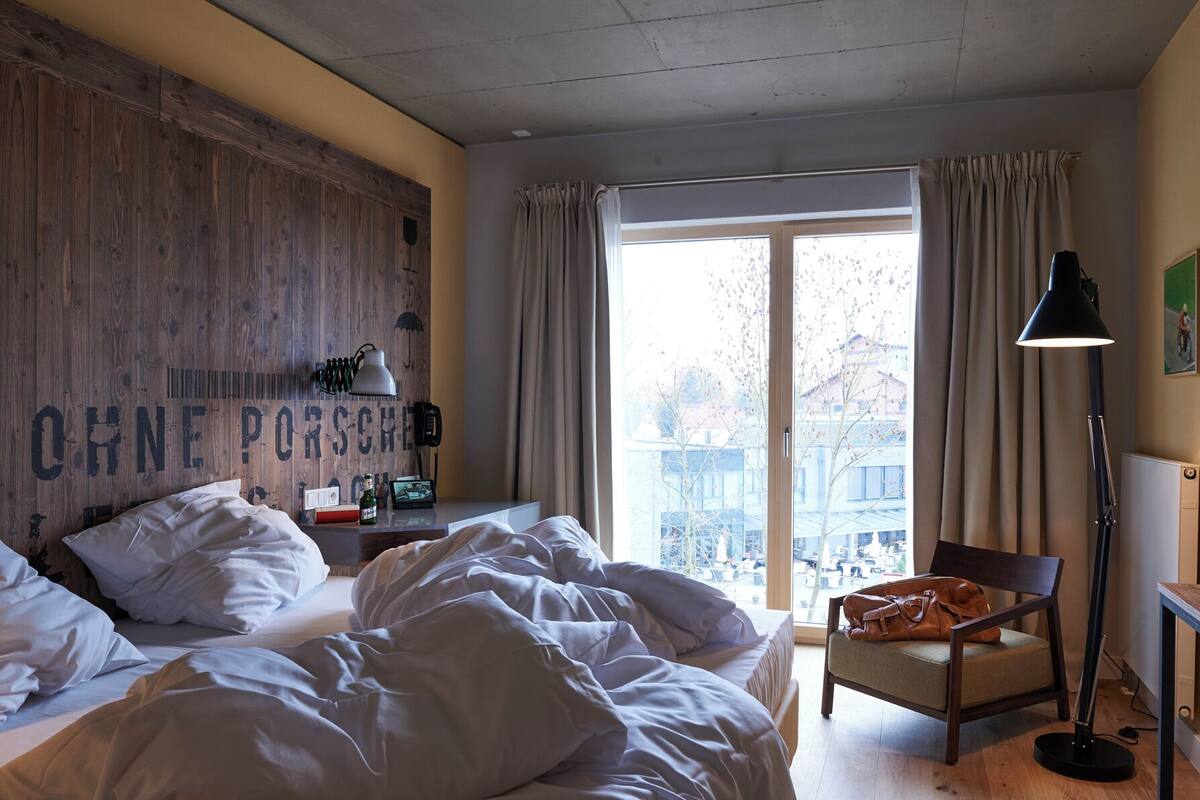 A bedroom with a bed, a chair, a desk with a lamp, a window with curtains, and a wall with a mounted phone and lamp.