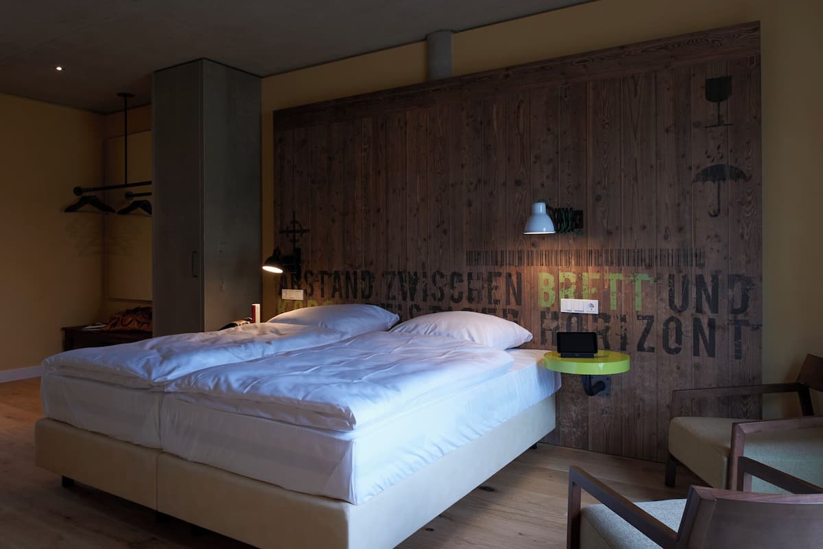 A bedroom with a large bed, a wooden accent wall, a bedside table with a lamp, and a chair.
