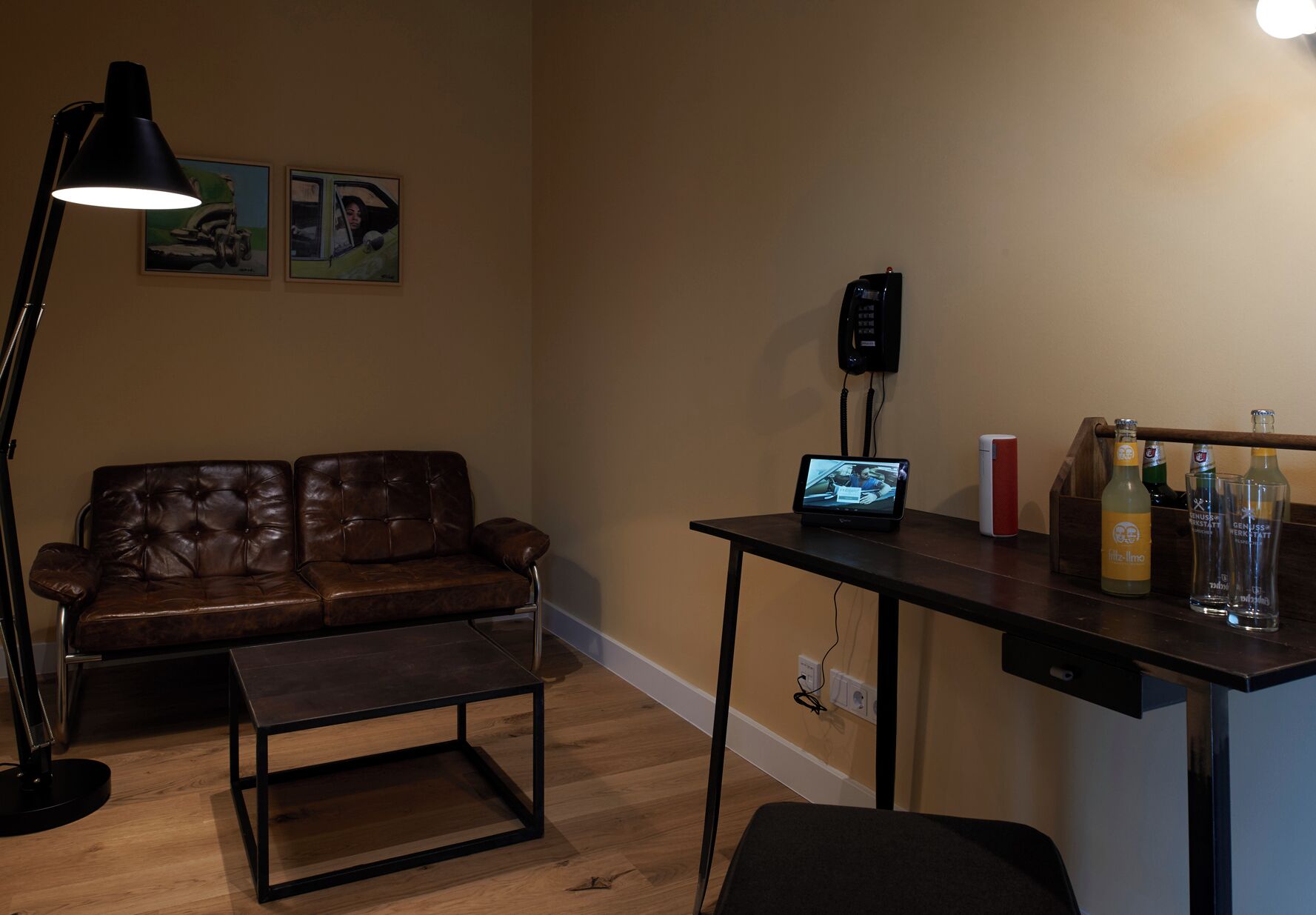 Een woonkamer met een bruine leren bank, een bijzettafeltje, een bureau met een computer, een lamp en een aan de muur bevestigd telefoon.