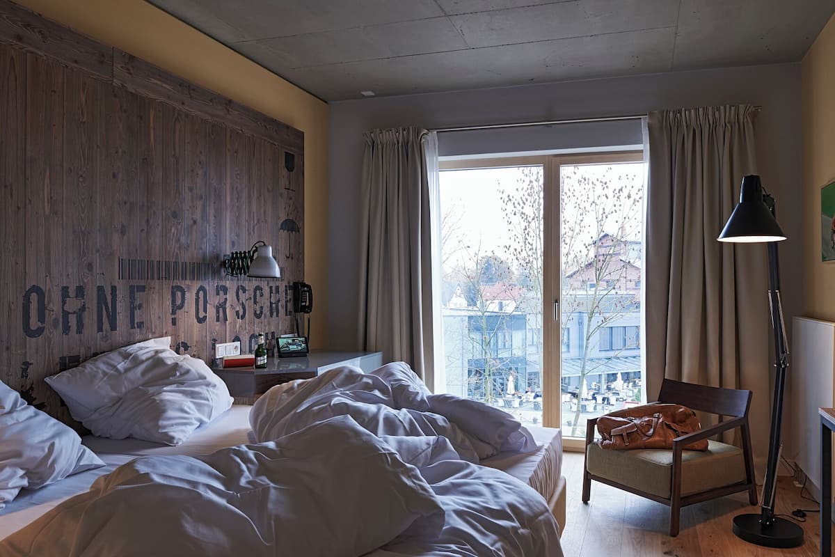 A hotel room with a bed, a desk with a lamp, a chair, and a large window with curtains.