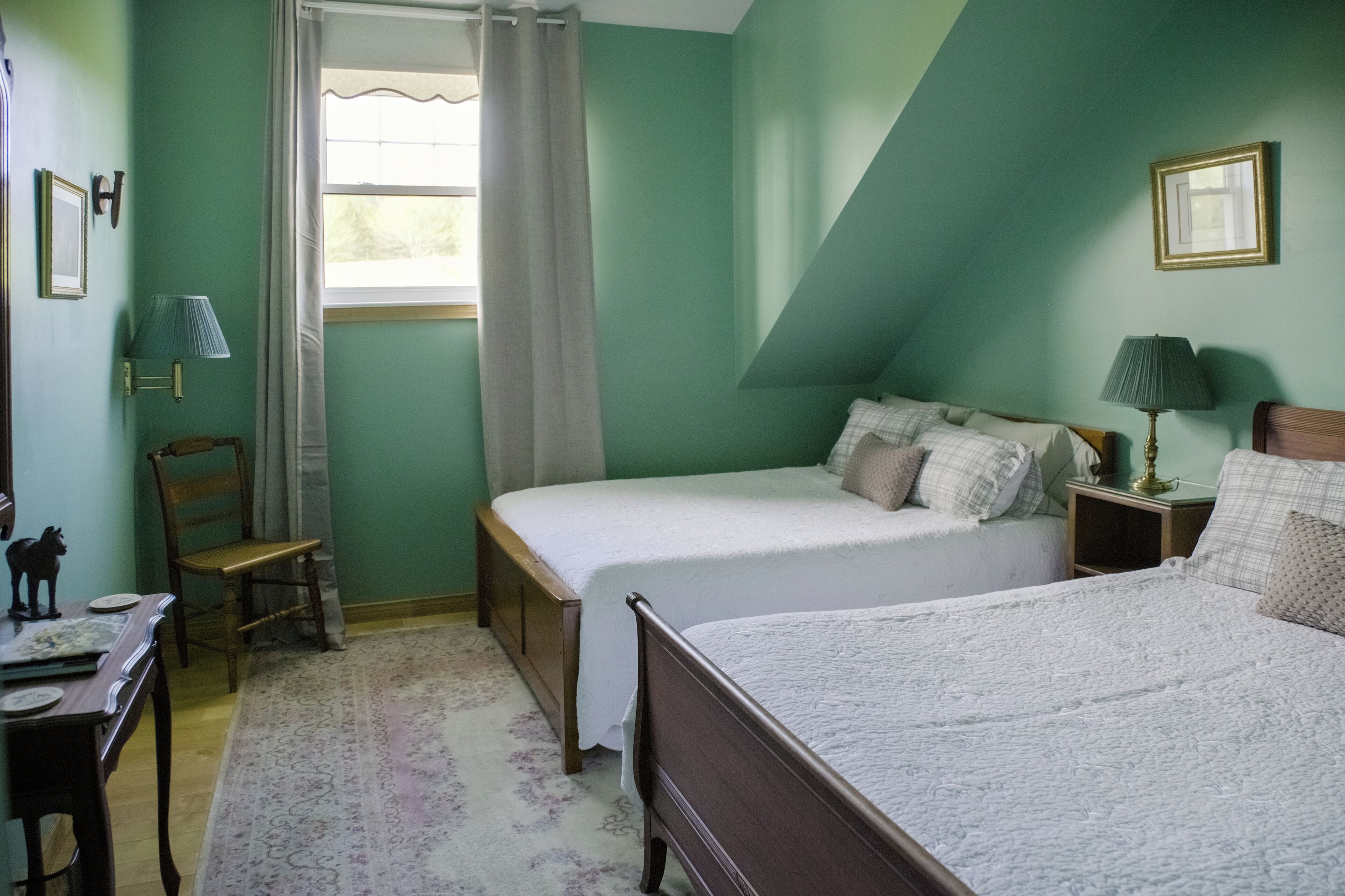 A bedroom with two beds, a window with curtains, a wooden chair, a small table, and a lamp.
