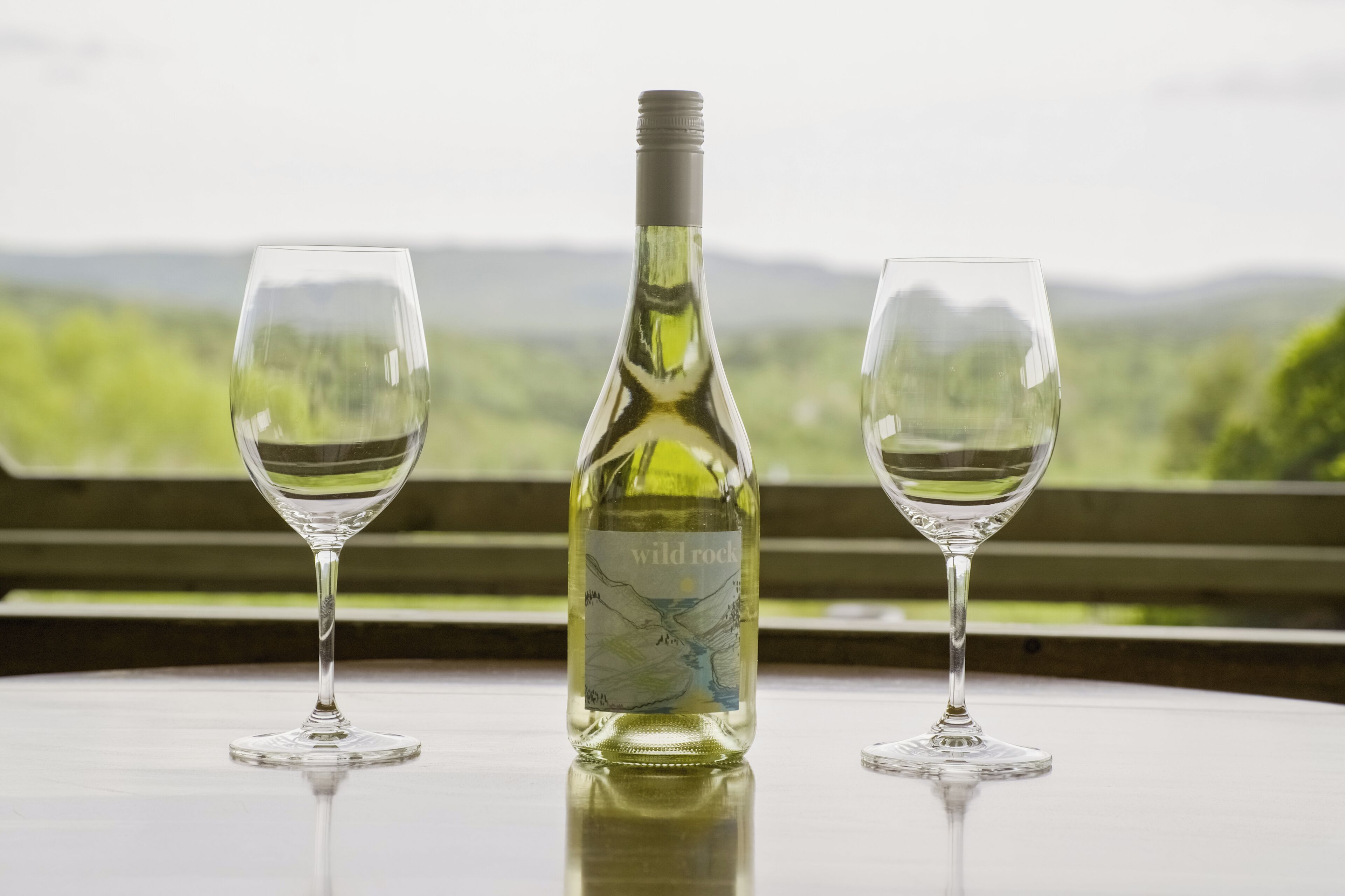 A bottle of wine and two glasses on a table with a view of green hills in the background.