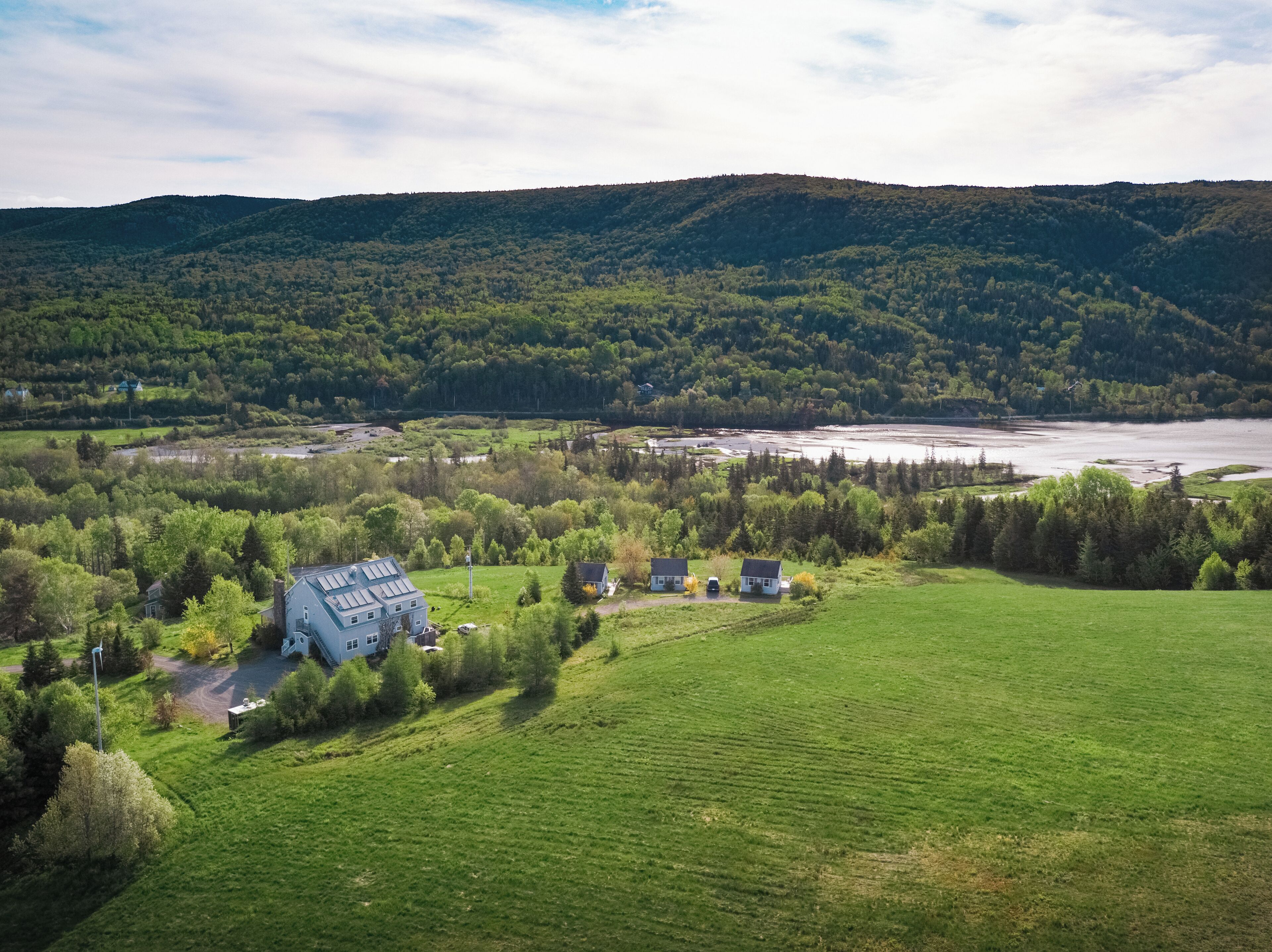 A large house with a sloped roof and multiple windows, surrounded by lush green fields and a river in the background.