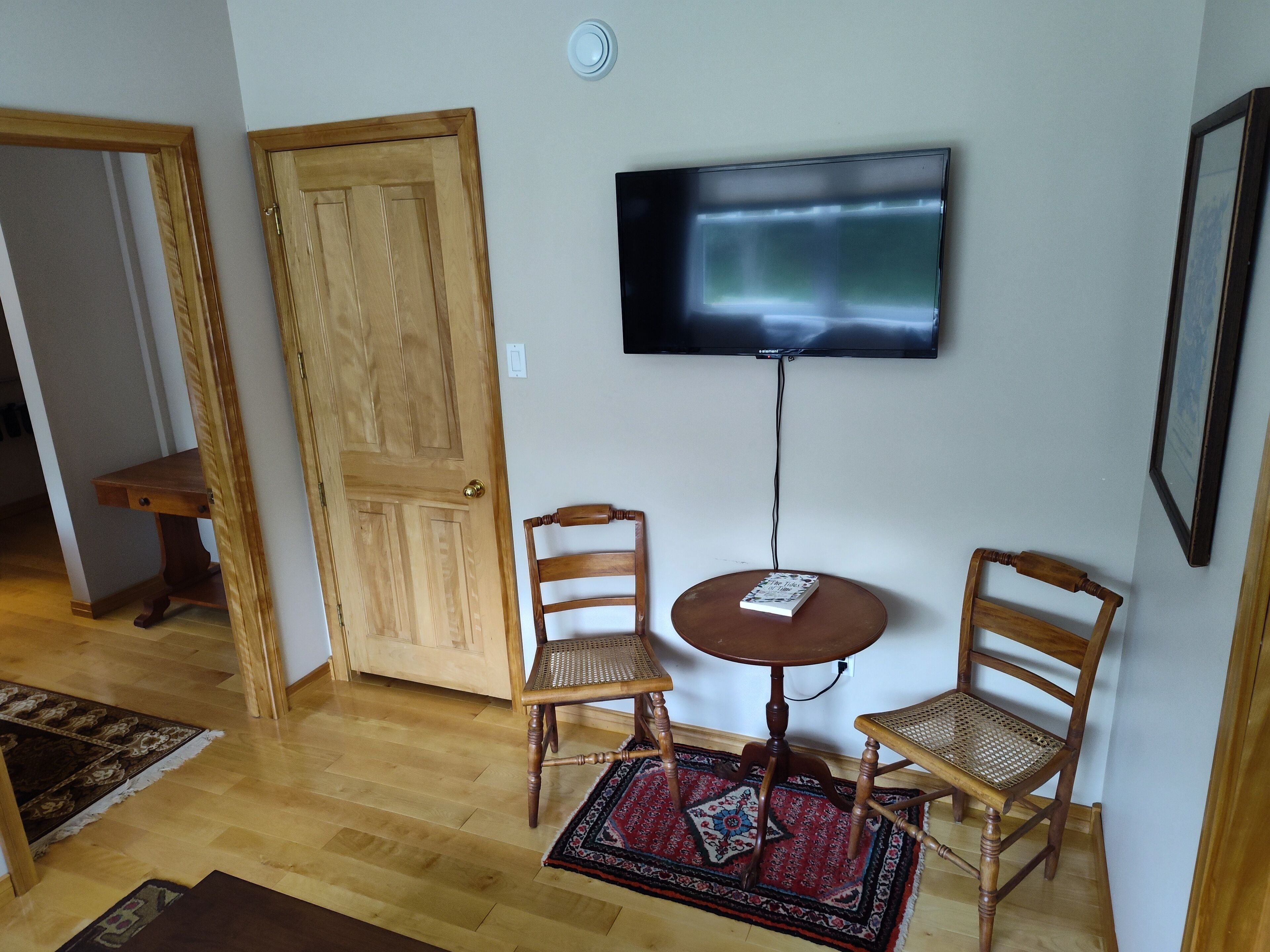 Een kamer met een houten deur, een aan de muur bevestigde televisie, twee houten stoelen, een klein rond tafeltje en een kamerkleed met een patroon op de vloer.