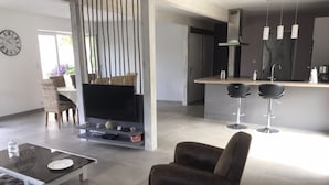 A modern living room with a flat-screen TV, a coffee table, and a sofa. There is a kitchen area with a bar and two bar stools.