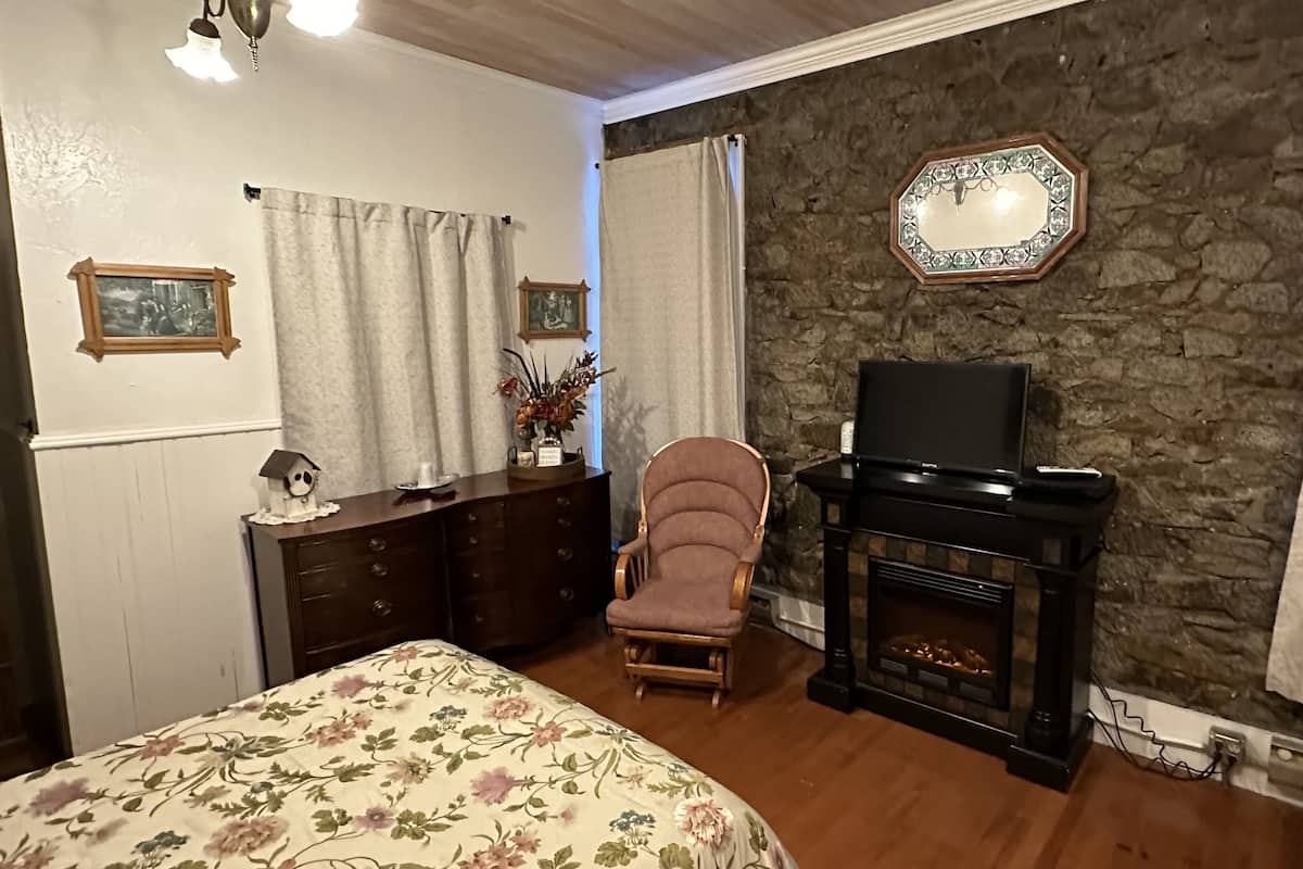 A bedroom with a bed, dresser, chair, television, fireplace, and stone wall.