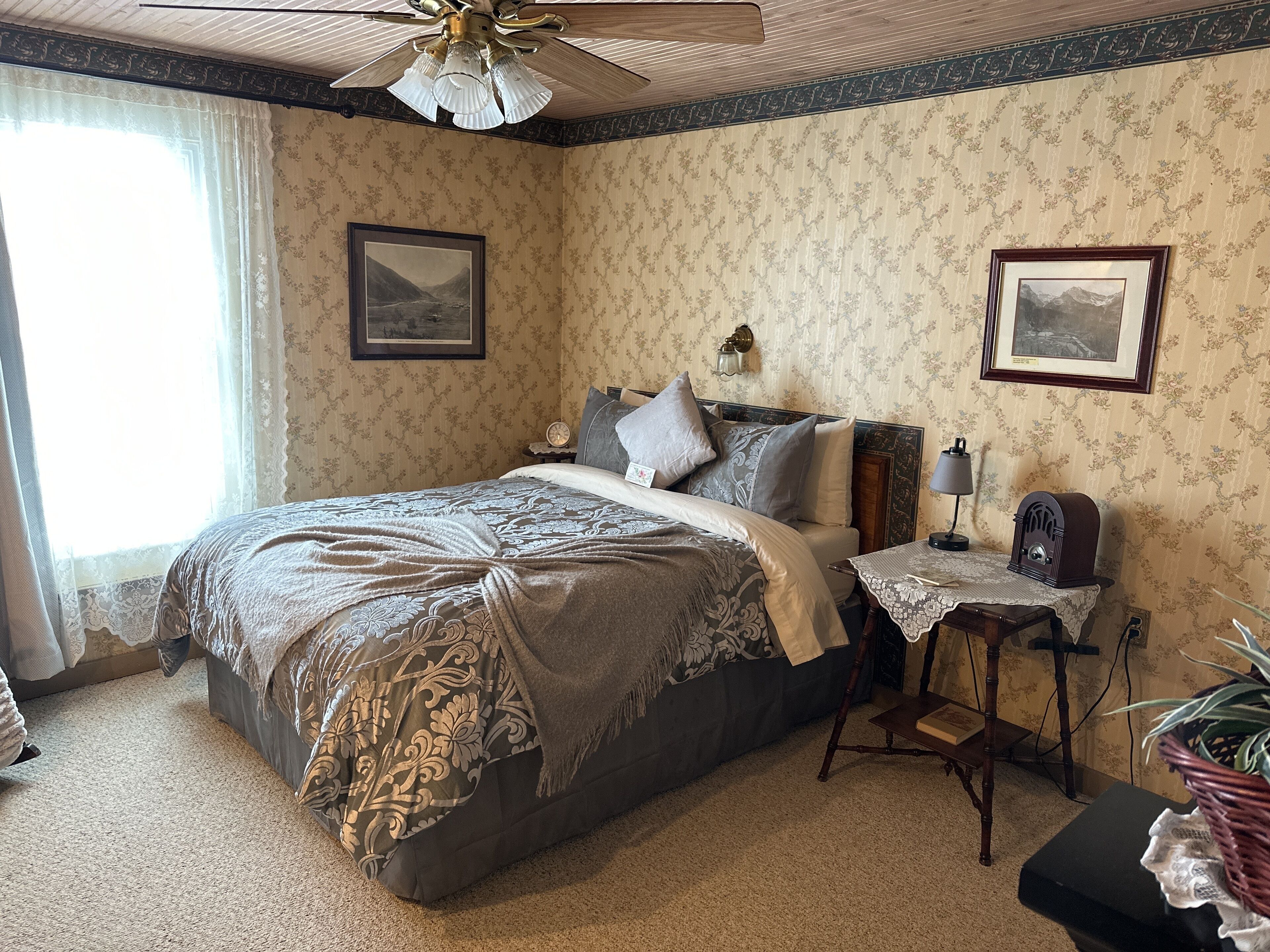 A bedroom with a large bed, a ceiling fan, a nightstand with a lamp, and two framed pictures on the wall.