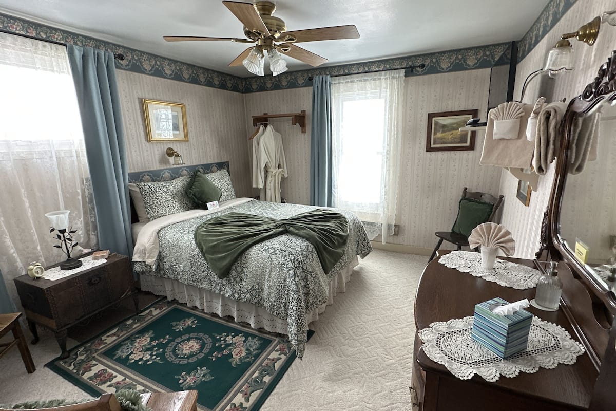 A bedroom with a bed, a dresser, a chair, a nightstand, a ceiling fan, and a window with curtains.